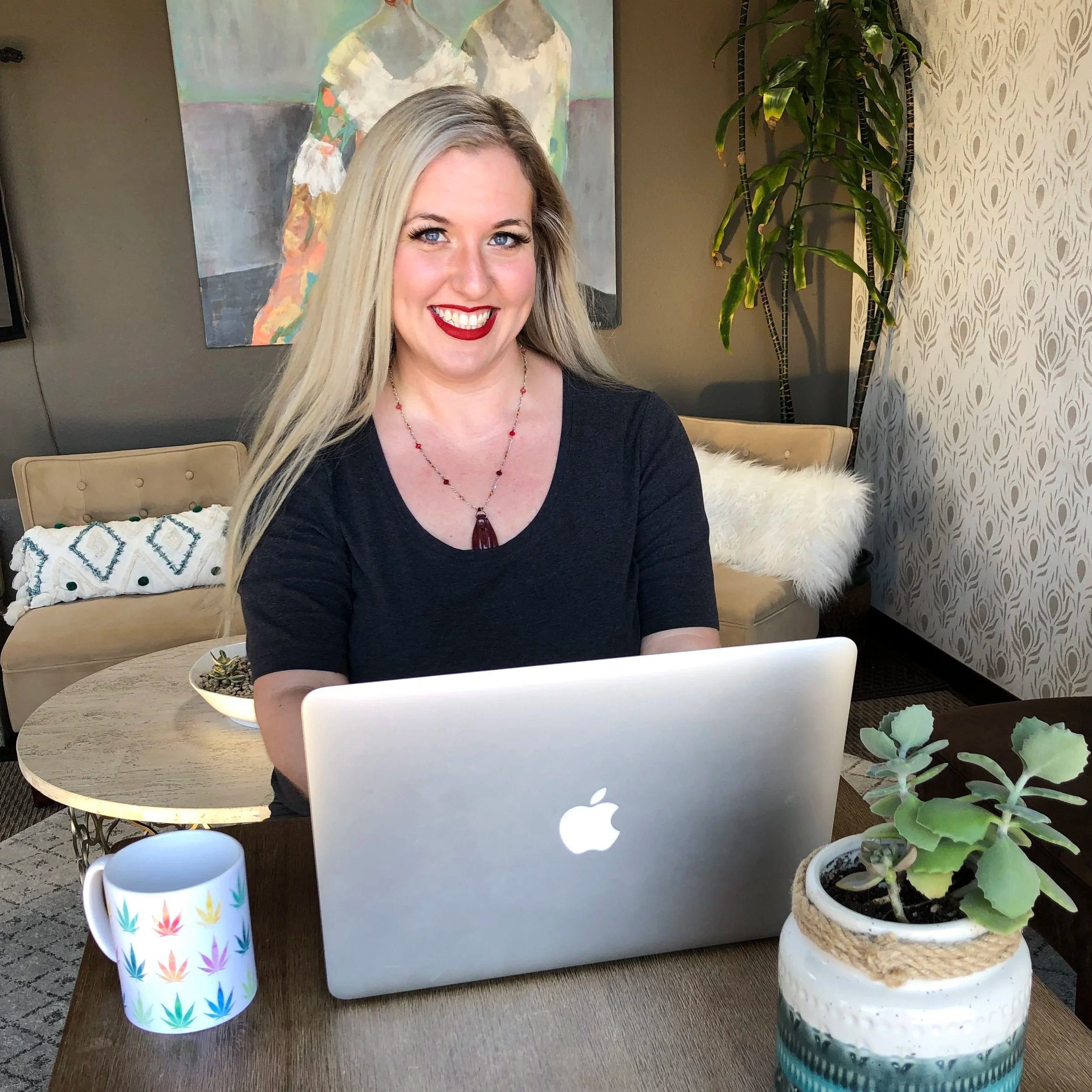 A woman with long blonde hair, wearing red lipstick and a black top, sitting at a table with a silver laptop in front of her. There are three potted plants on the table, one of which has succulents, and a colorful mug with a leaf pattern. A painting of two women with their backs turned is on the wall behind her, along with a tall green plant and decorative wallpaper.