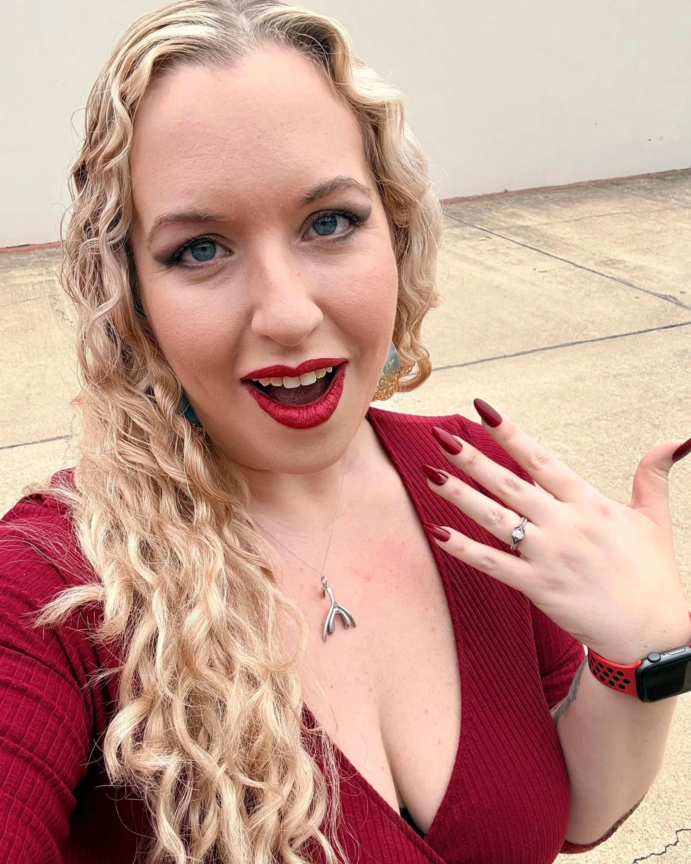 A woman with curly blonde hair, wearing red lipstick, a red top, earrings, a ring, and a smartwatch, taking a selfie outdoors on a concrete sidewalk with a beige wall background.
