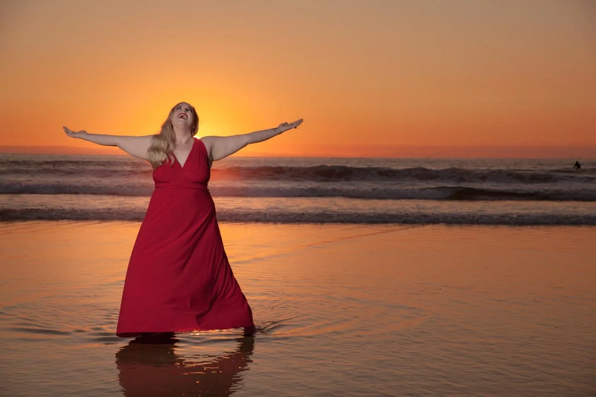 Woman in a red dress standing in the shallow water on the beach at sunset with arms outstretched, smiling and looking up.