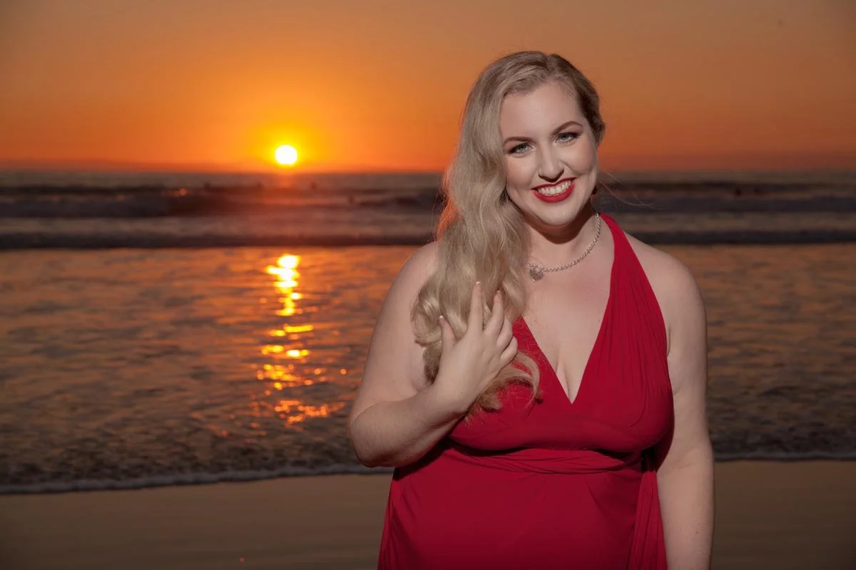 A woman with long blonde hair wearing a red dress smiling at the camera on a beach at sunset