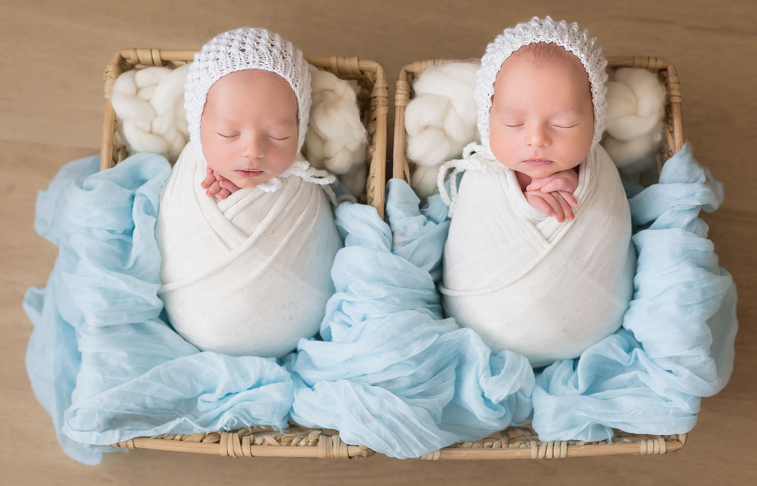Two sleeping newborn babies wrapped in white blankets and wearing knitted caps, lying in a wicker basket with soft blankets, viewed from above.
