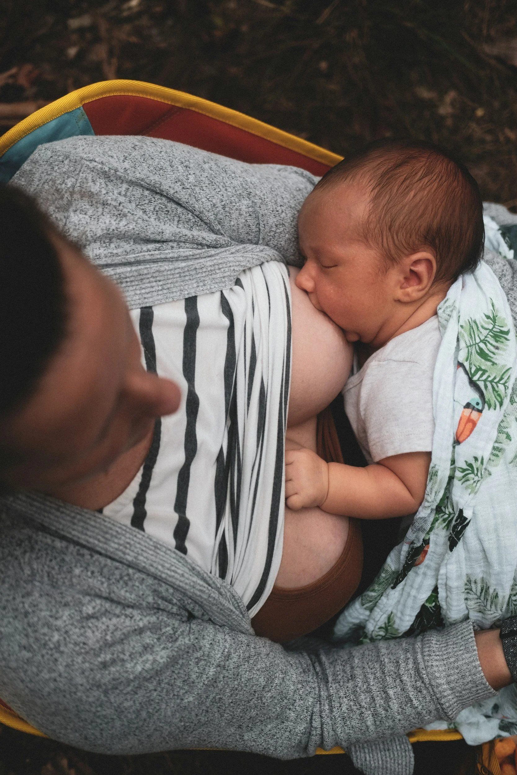 A baby breastfeeding on a person wearing a gray sweater and striped shirt, lying on a yellow and red surface outdoors with wood chips on the ground.