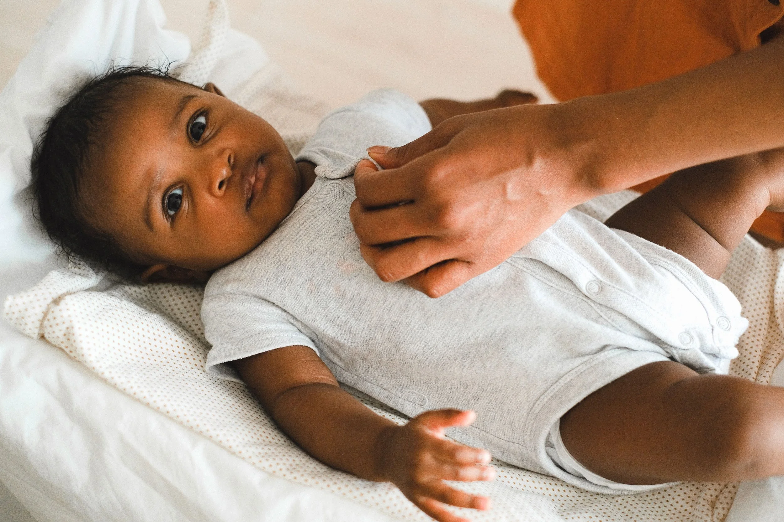 A young child lying in a bed, looking at the camera, with an adult hand gently holding their arm.