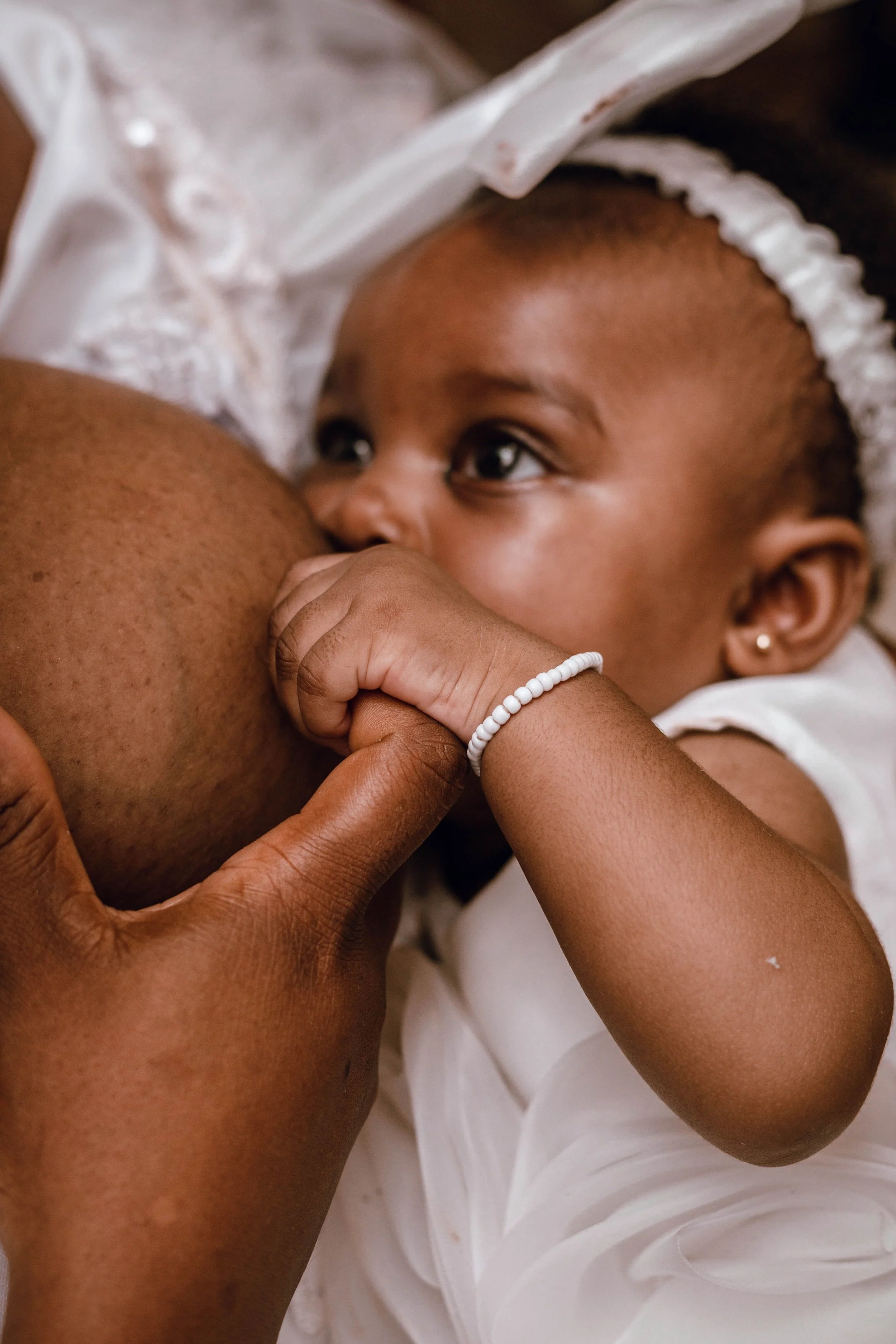 A young child breastfeeding, gently holding her mother's breast and wearing a white beaded bracelet and a white cap.