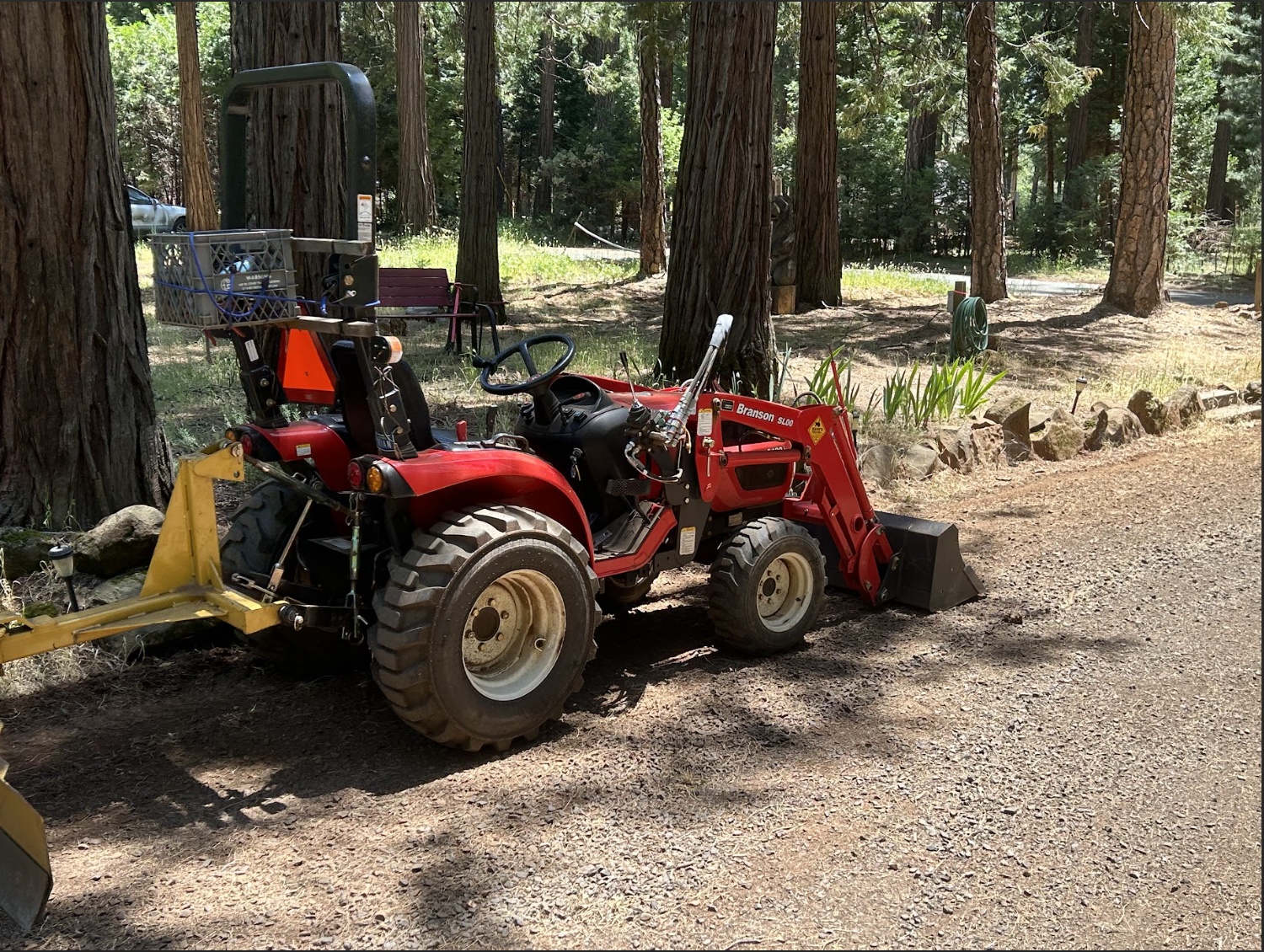 A red tractor with front loader attachment parked on a dirt pathway in a wooded area with tall trees and some green plants.