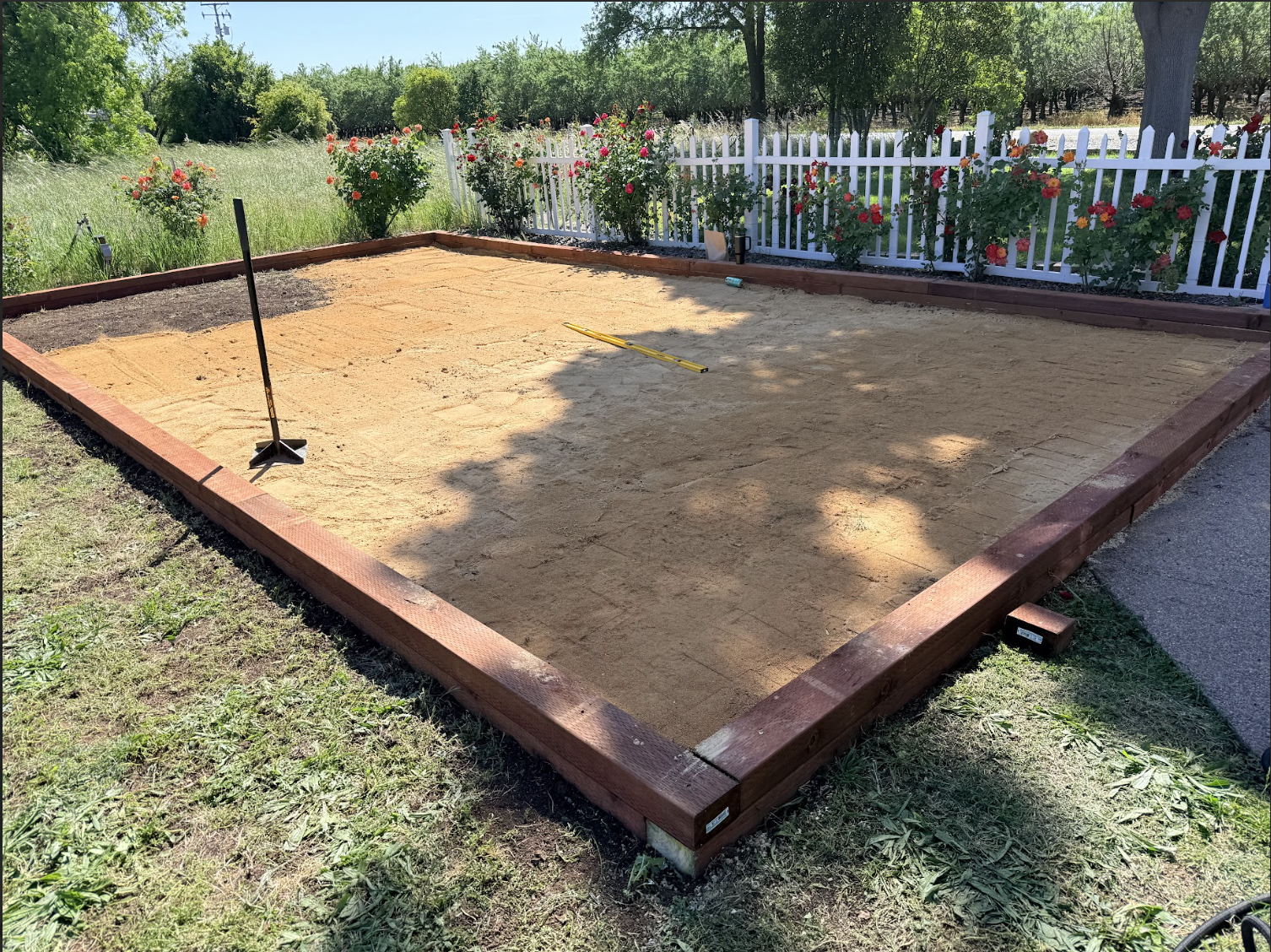 A backyard with a new wooden deck being built, with sand spread on top and construction tools like a level and rake on the ground, surrounded by green grass, trees, and a white picket fence with flowering plants.