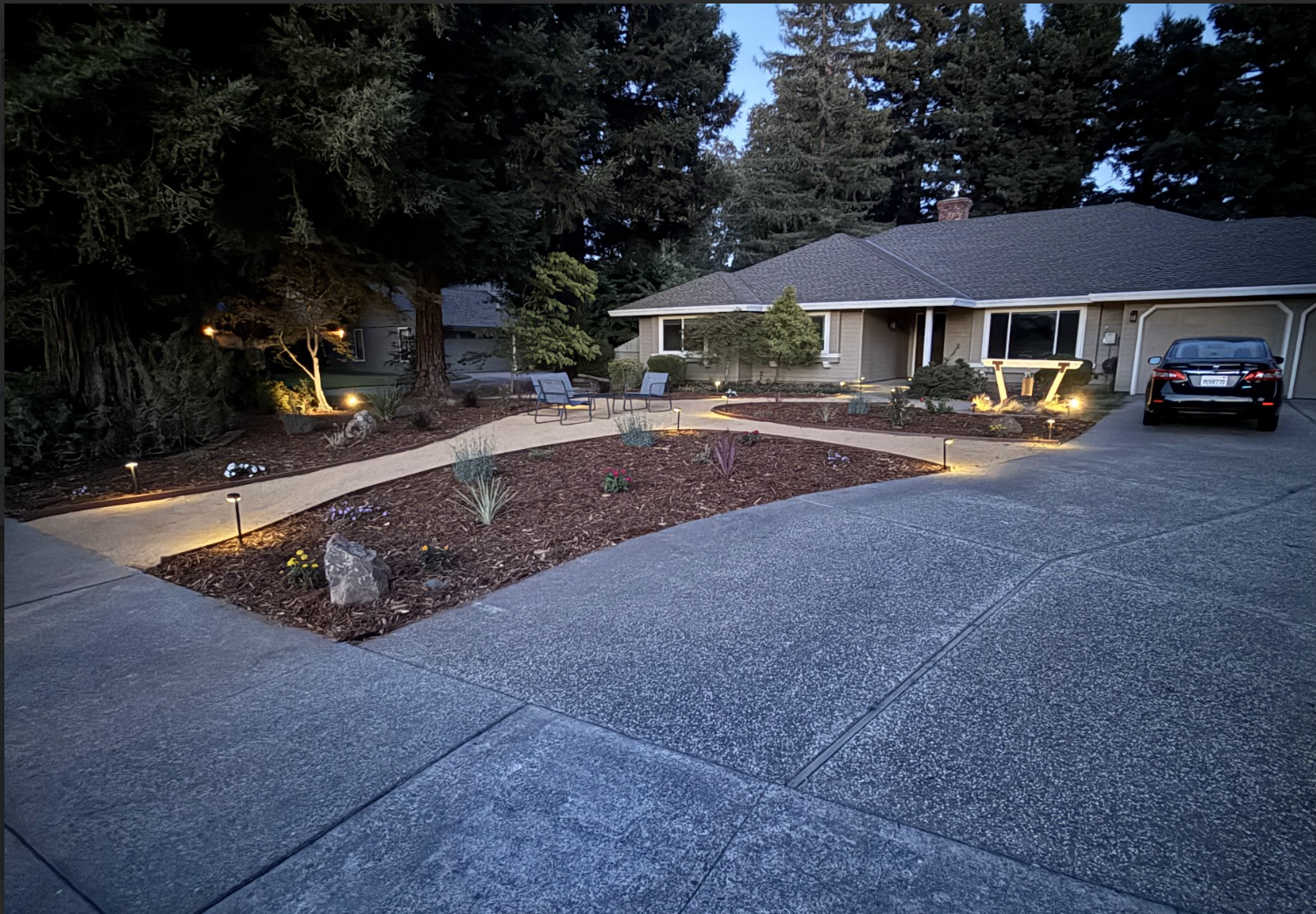 A well-lit front yard of a house during dusk with a concrete driveway, a landscaped garden with small plants, path lights, and outdoor chairs, with large trees and a house with a garage and parked car in the background.