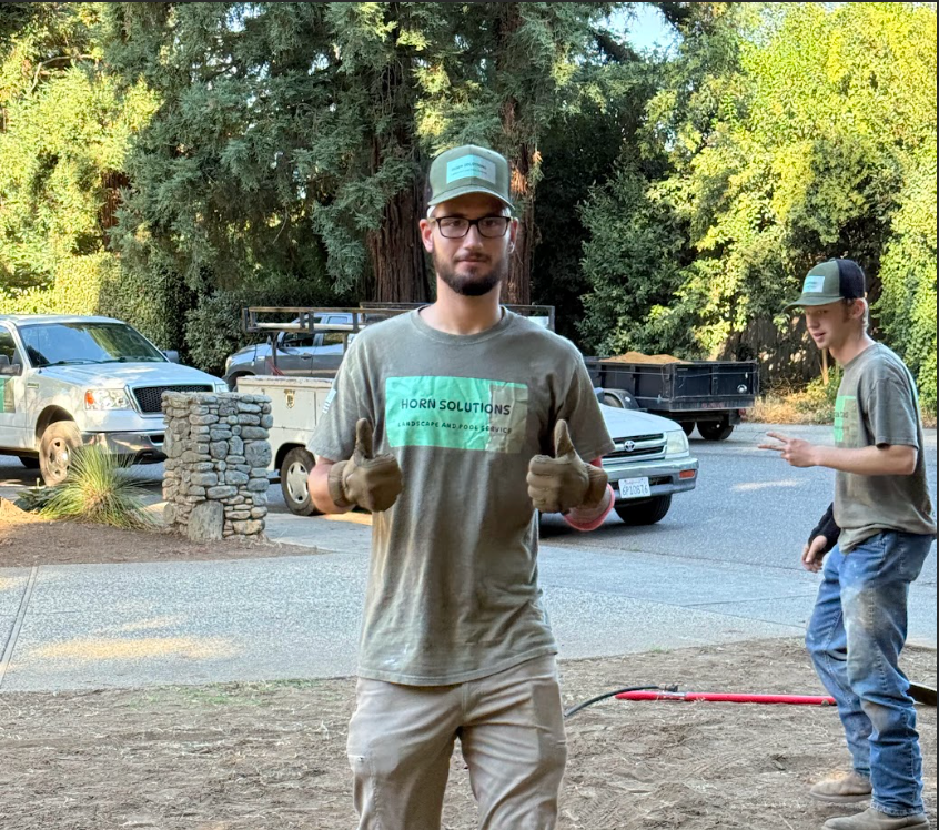 Young man wearing glasses, a cap, and gloves standing outdoors, giving two thumbs up in front of parked cars and green trees.
