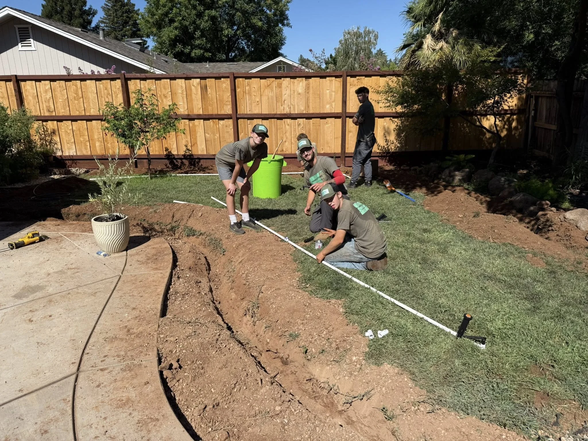 Group of young men working on landscaping or gardening project in backyard, installing part of a fence or border, with tools and a green trash bin nearby.