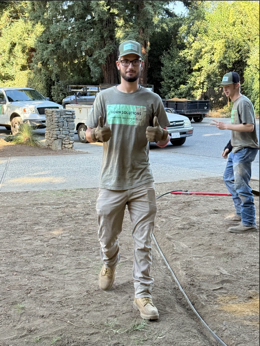 A man in work gloves and glasses giving a thumbs-up, standing outdoors on dirt, with trees, parked cars, and another person in the background.