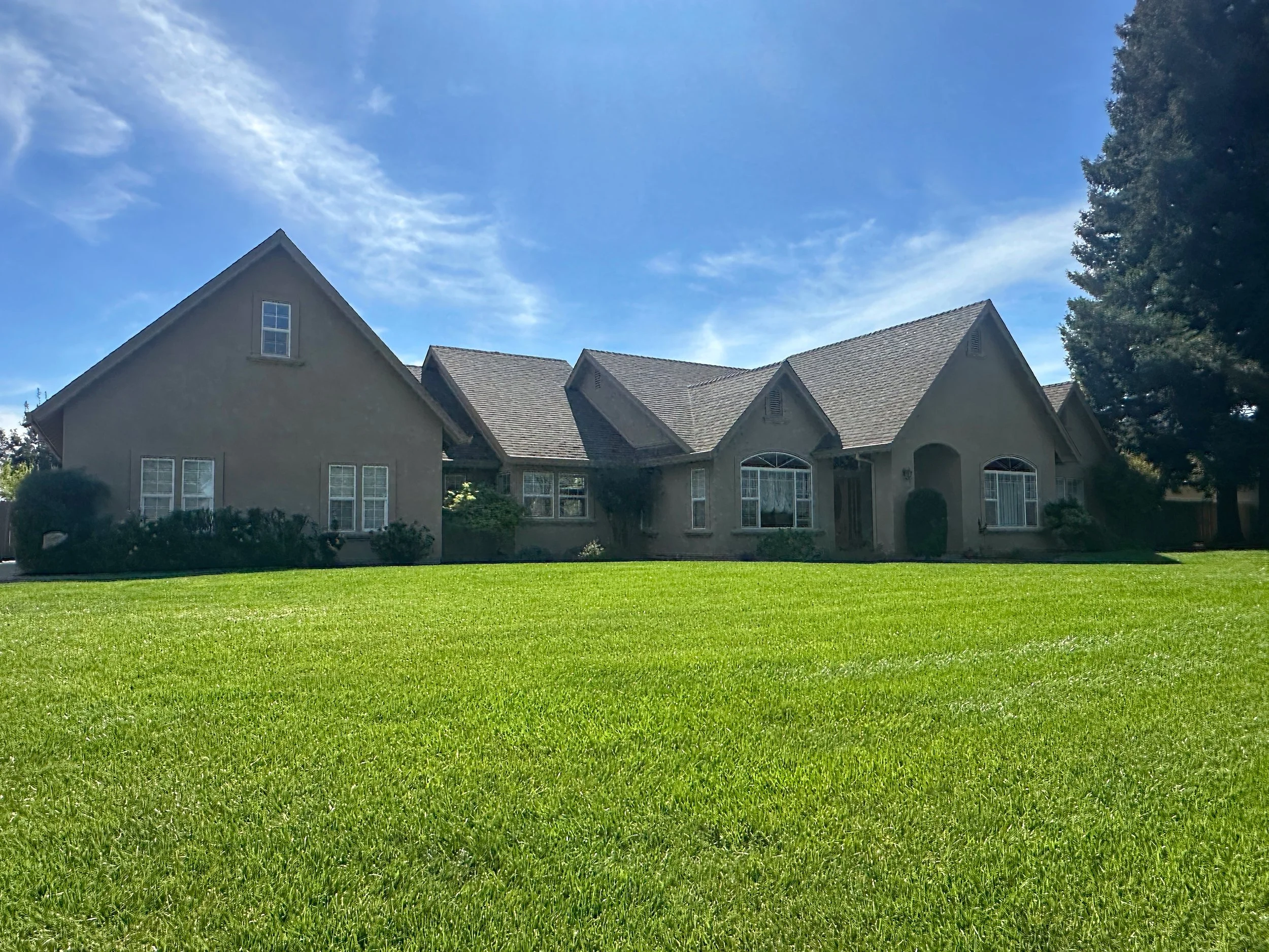 A large house with a brown roof and multiple gables, surrounded by a well-kept lawn and greenery, under a bright blue sky with some clouds.