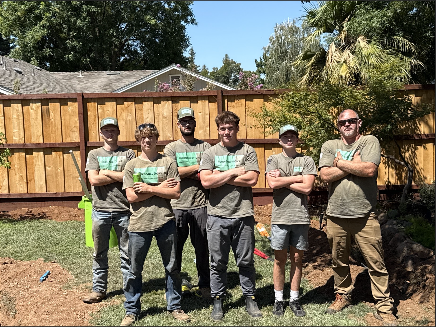 Group of six males standing outdoors in front of a wooden fence, with some working tools and plants visible, on a sunny day.