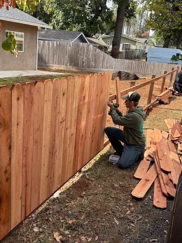 A man kneeling and using a drill to assemble a wooden fence in a backyard with trees and neighboring houses.