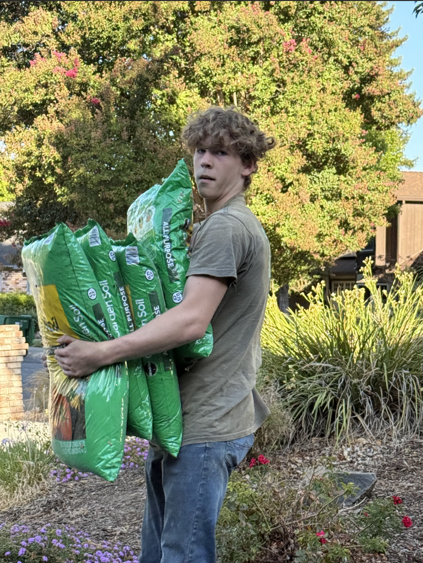 Young man holding multiple green bags of potting soil outdoors with trees and plants in the background.