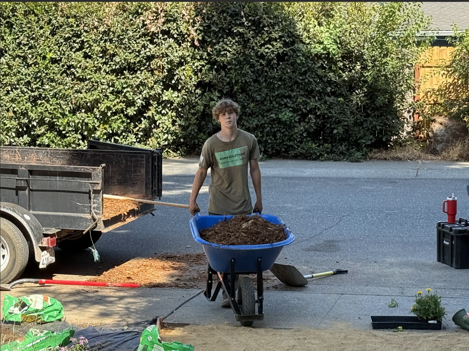 A young person with curly hair is pushing a blue wheelbarrow filled with mulch or soil in a residential driveway. There is a trailer with more mulch attached, and gardening supplies such as a shovel, a rake, and a bag of soil are on the ground. A bla