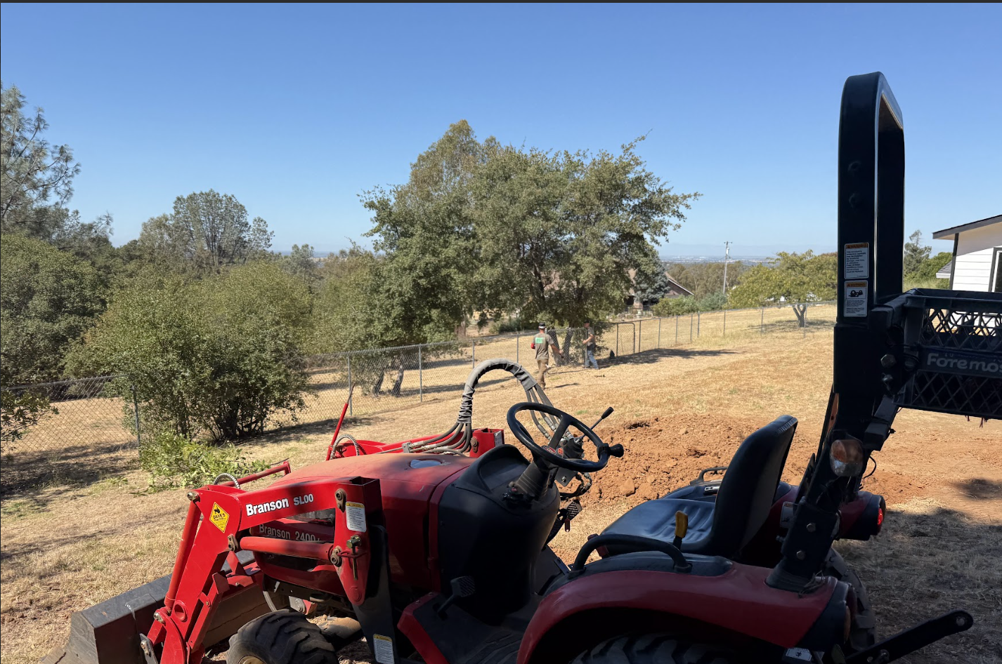 A red Branson mini tractor with a front loader in a backyard, with a chain-link fence, trees, and two people working in the distance