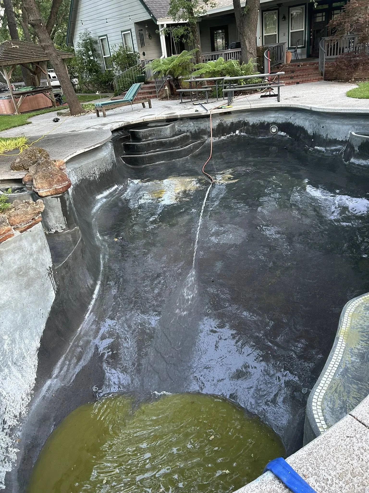 An empty in-ground swimming pool with a dark lining and a small section filled with dirty green water, surrounded by a concrete deck with steps leading into the pool. In the background, there is a house with a porch, trees, outdoor furniture, and gar