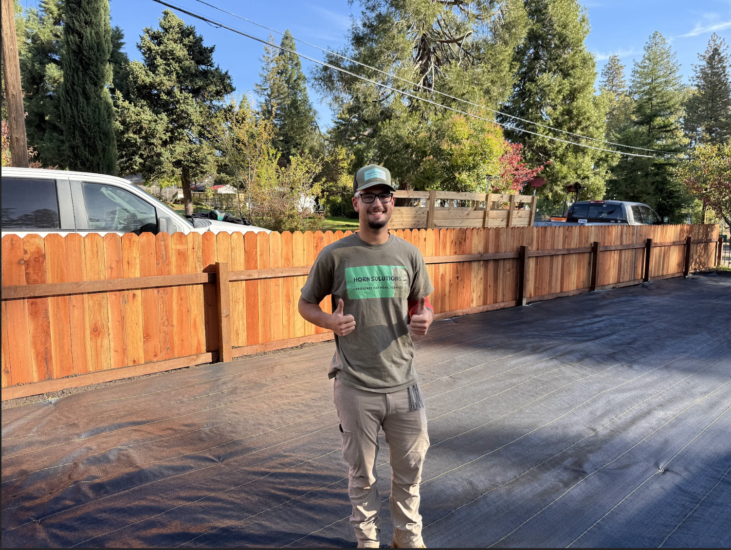 A man stands in front of a wooden fence, smiling and giving two thumbs up, with cars and trees in the background on a sunny day.