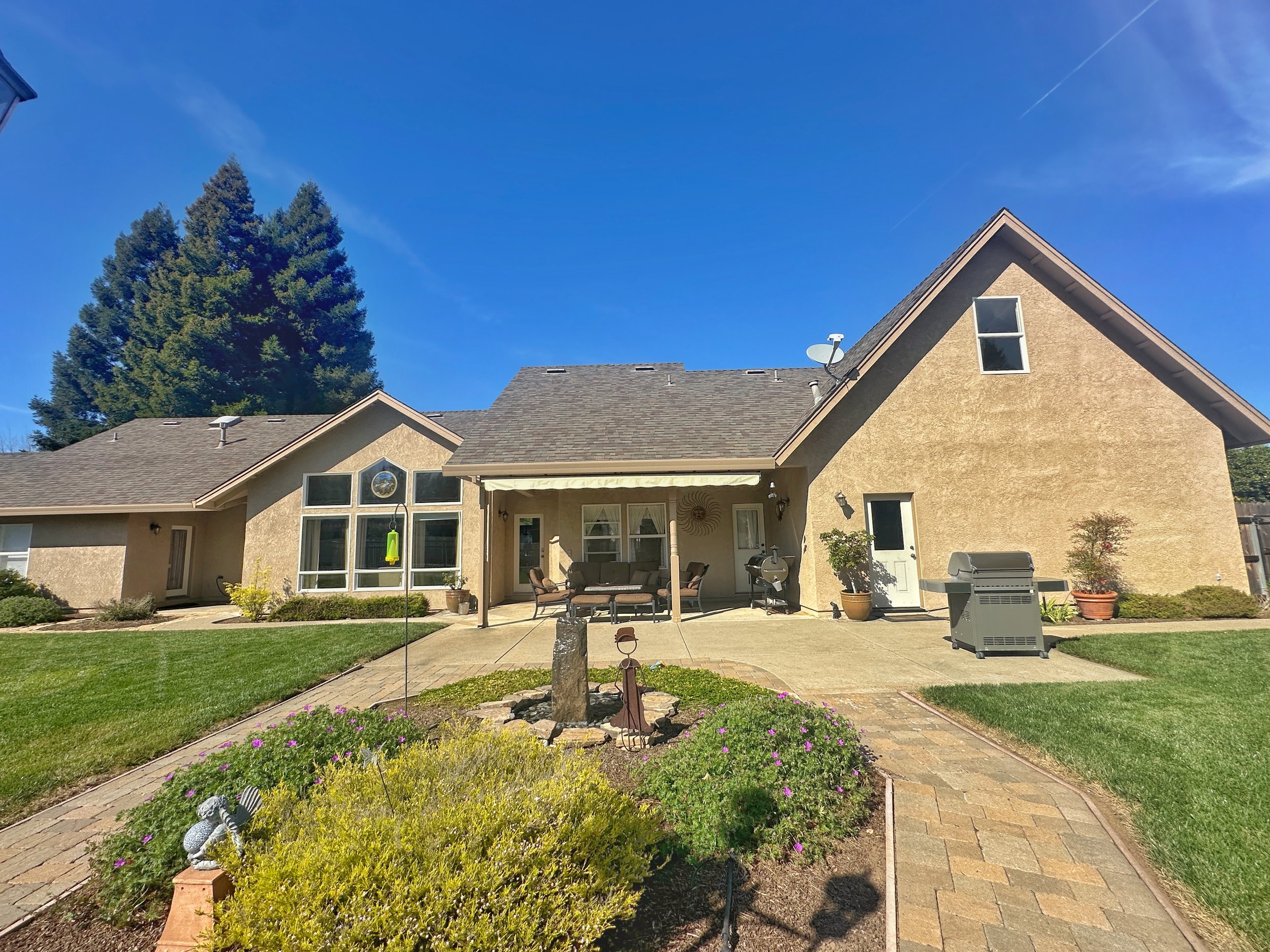 A backyard patio scene with a cushioned sofa set under a roofed patio area, a barbecue grill, potted plants, and a small garden with purple flowers and decorative garden ornaments, surrounded by a well-maintained lawn, with the house in the backgroun