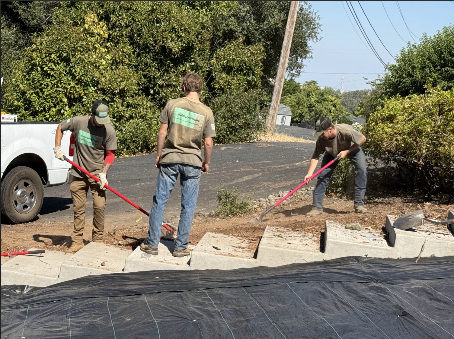 Three men working outdoors on sidewalk construction, using shovels to move dirt near concrete blocks, with a white pickup truck and green trees in the background.