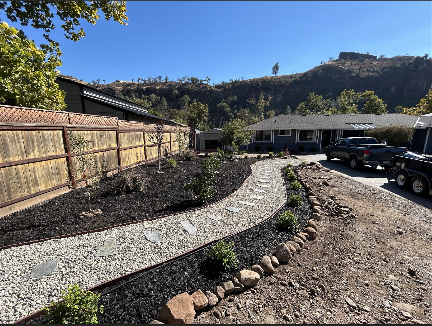 A landscaped front yard with a curved gravel pathway, small bushes, and young trees, bordered by rocks, in front of a house with a mountain backdrop, blue sky, and parked trucks.