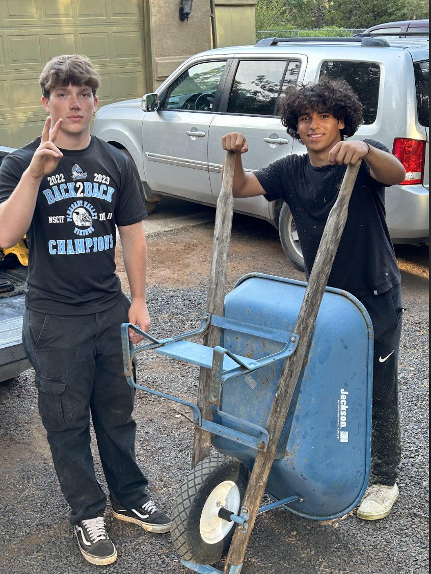 Two young men pose with a blue wheelbarrow outdoors in front of a silver SUV. The young man on the left is making a peace sign with his fingers and wearing a black T-shirt with the words 'Back 2 Back Champions' and a logo. The young man on the right 