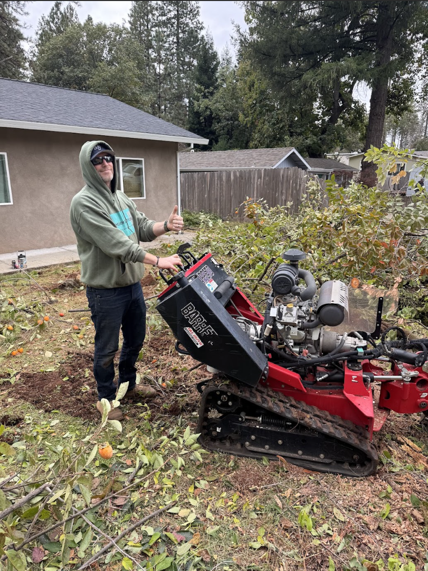 A person wearing sunglasses, a hoodie, and jeans, giving a thumbs-up and operating a mini tracked excavator in a backyard with fallen branches and orange fruit on the ground.