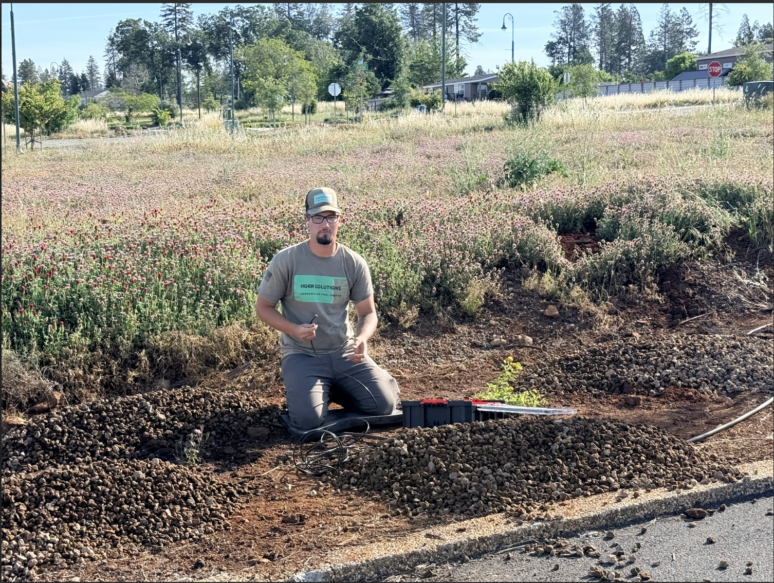 A man kneeling in a dirt area beside a roadside, holding wires and surrounded by tools, with a background of greenery,flowering plants, and trees.