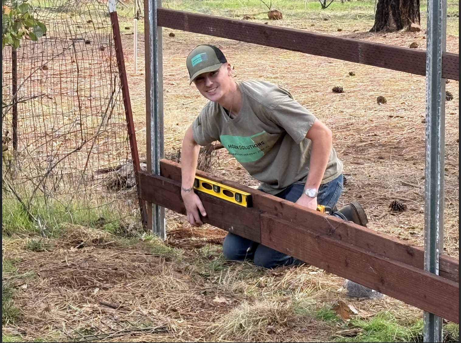 A man kneeling on the ground and working on a wooden fence with a level tool, outdoors with trees and dirt ground.