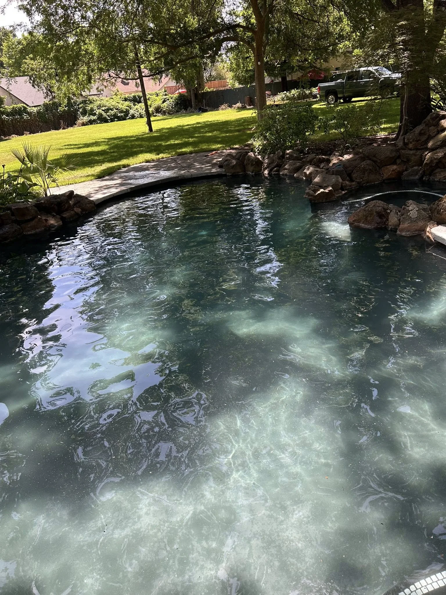 A backyard with a swimming pool, surrounded by a rock border and lush greenery, with trees providing shade and a truck parked on the grass.