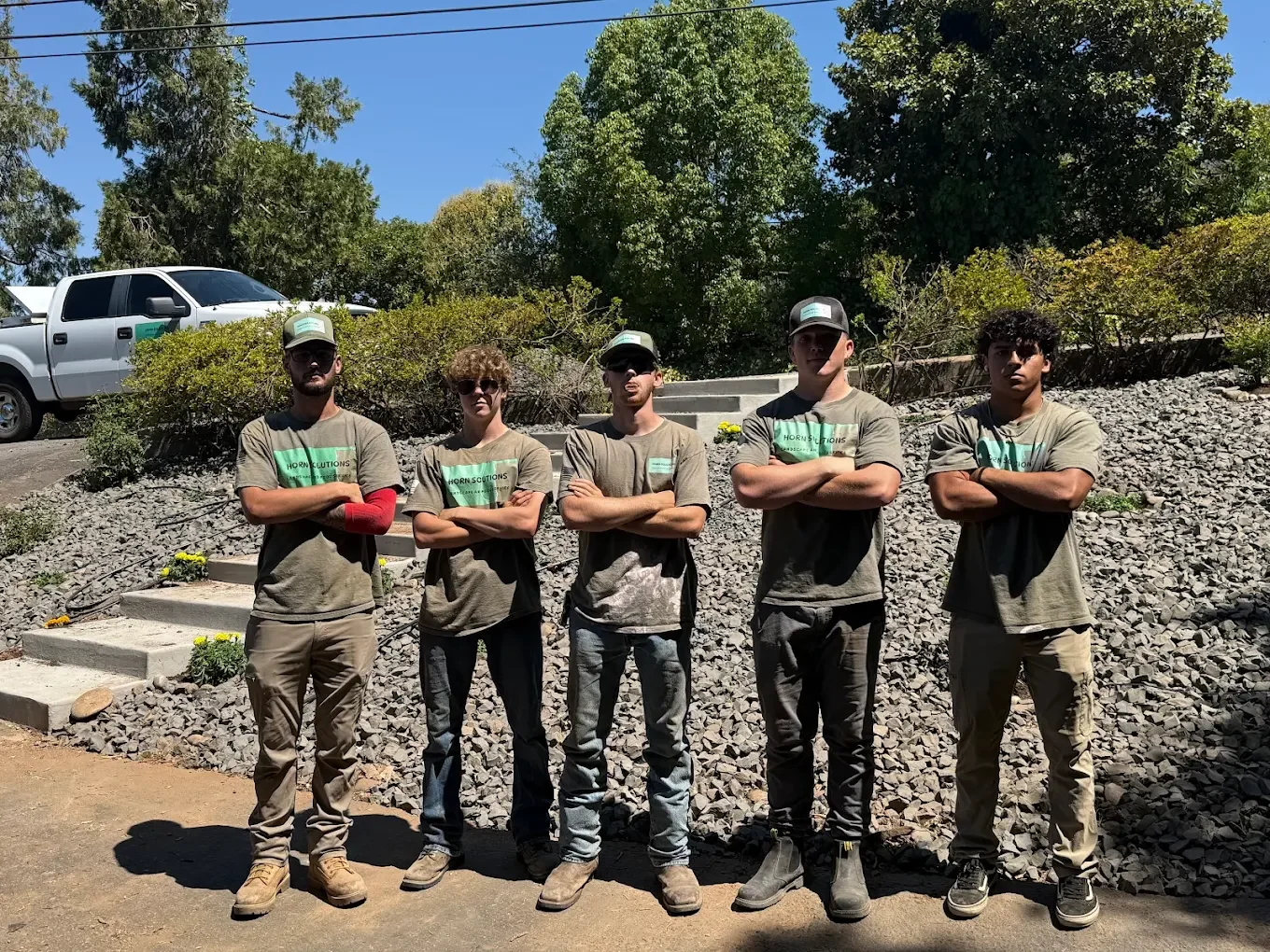 Five young men standing outdoors on a sunny day with trees and a parked truck in the background, all wearing matching gray t-shirts and caps with arms crossed.