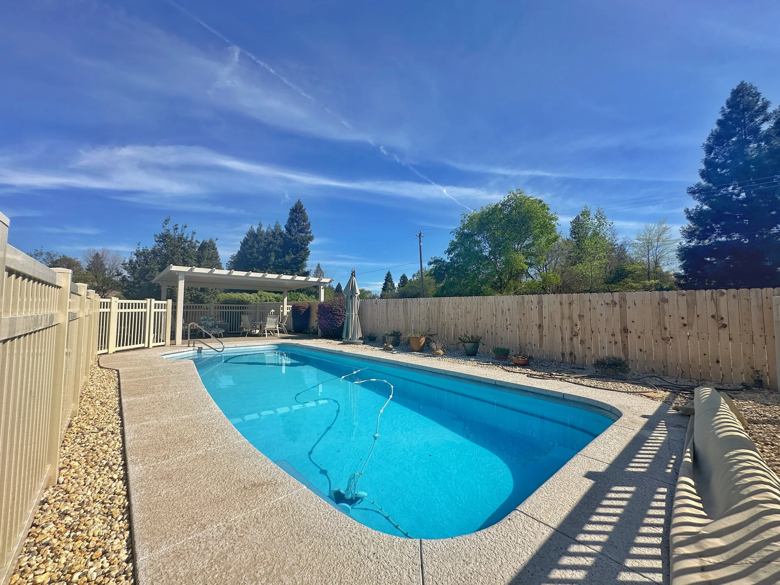 Backyard swimming pool with blue water, surrounded by concrete decking, a beige privacy fence, and a white pergola. There are trees and a clear blue sky with a few wispy clouds overhead.