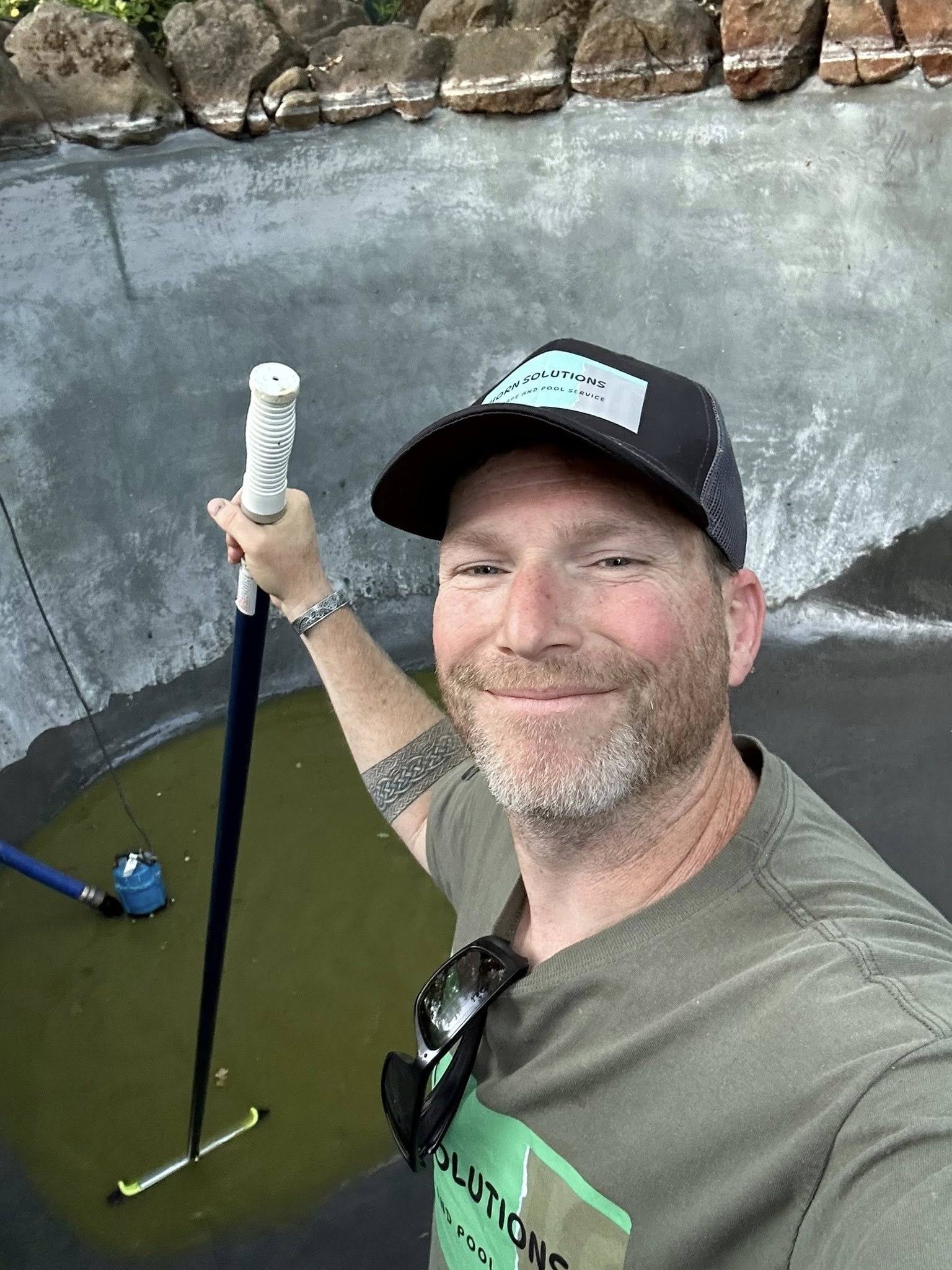 A man taking a selfie in a pool, holding a pool skimmer with a long handle. He is wearing a black cap with a logo, sunglasses hanging from his shirt, and a gray T-shirt with a green sign that reads 'SOLUTIONS POOL'.