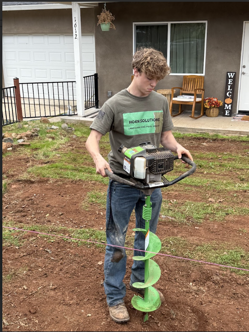 Young person using a green gasoline-powered earth auger on a patch of dirt in a backyard, with a house and porch in the background.