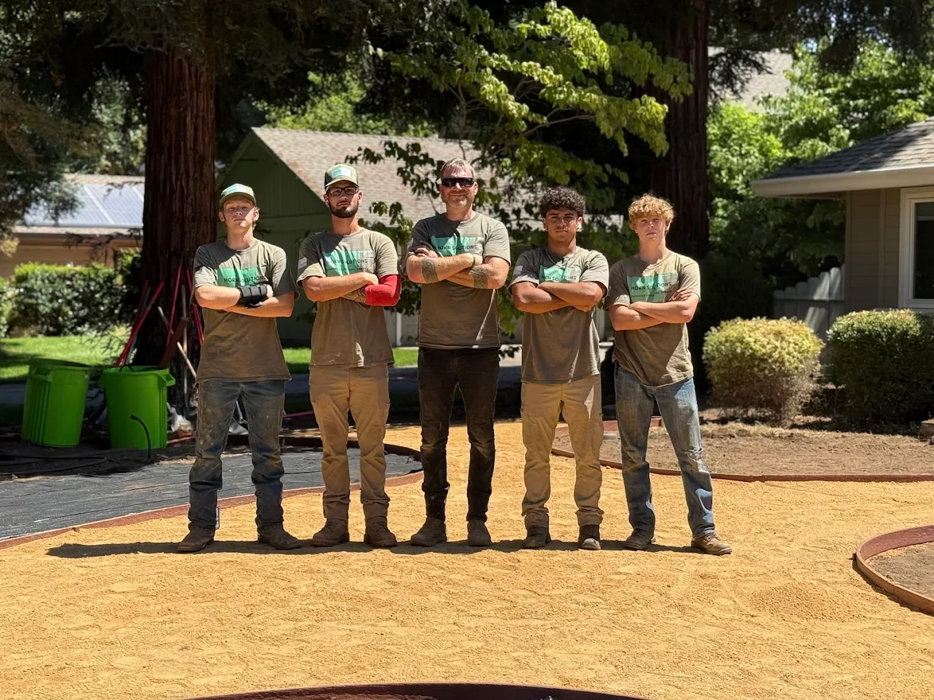 Five young men standing outdoors on a construction site with arms crossed, wearing matching grey t-shirts and jeans, surrounded by trees and houses.
