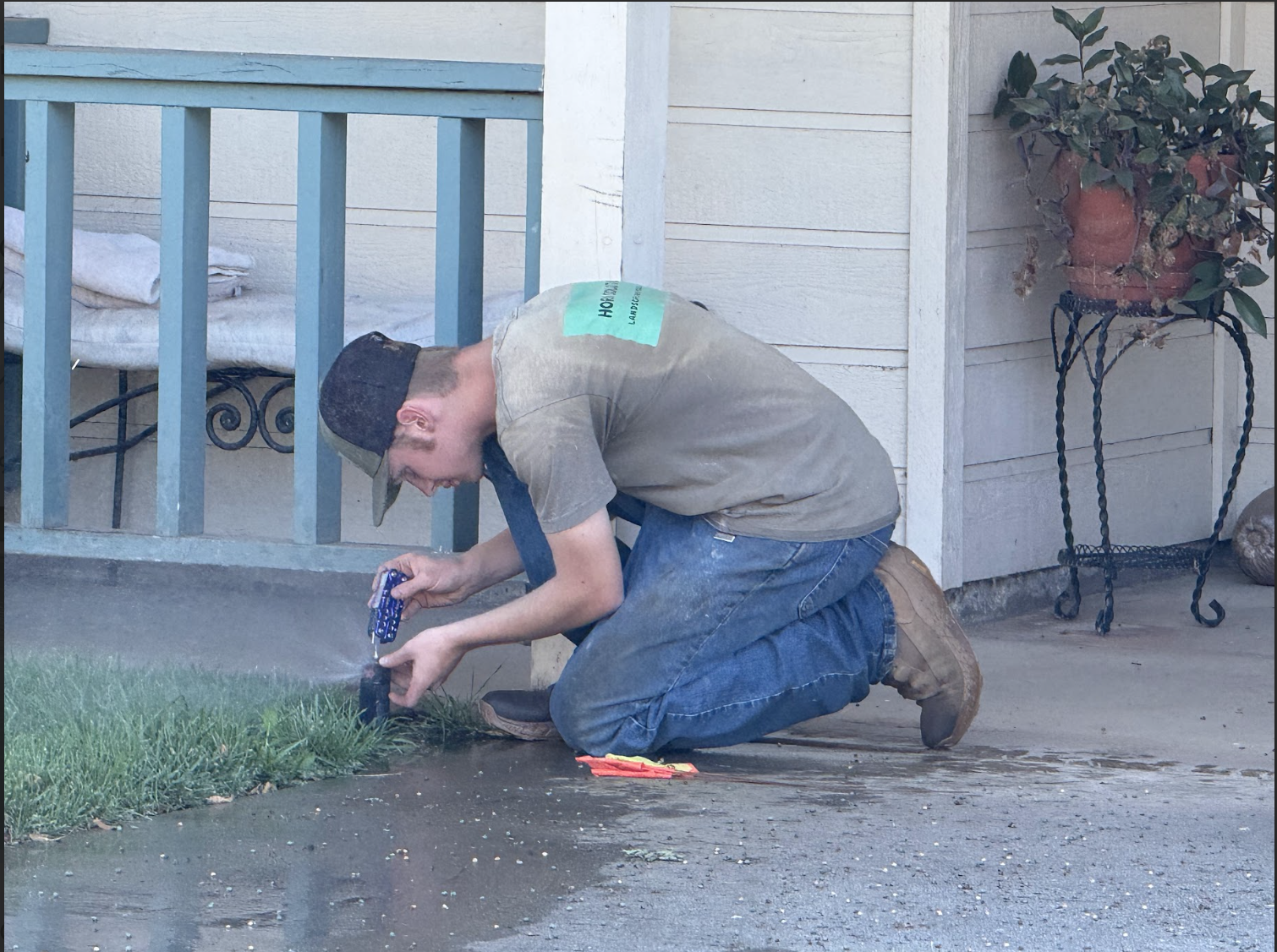 A man kneeling on the ground, watering the plants in a garden bed outside a house.
