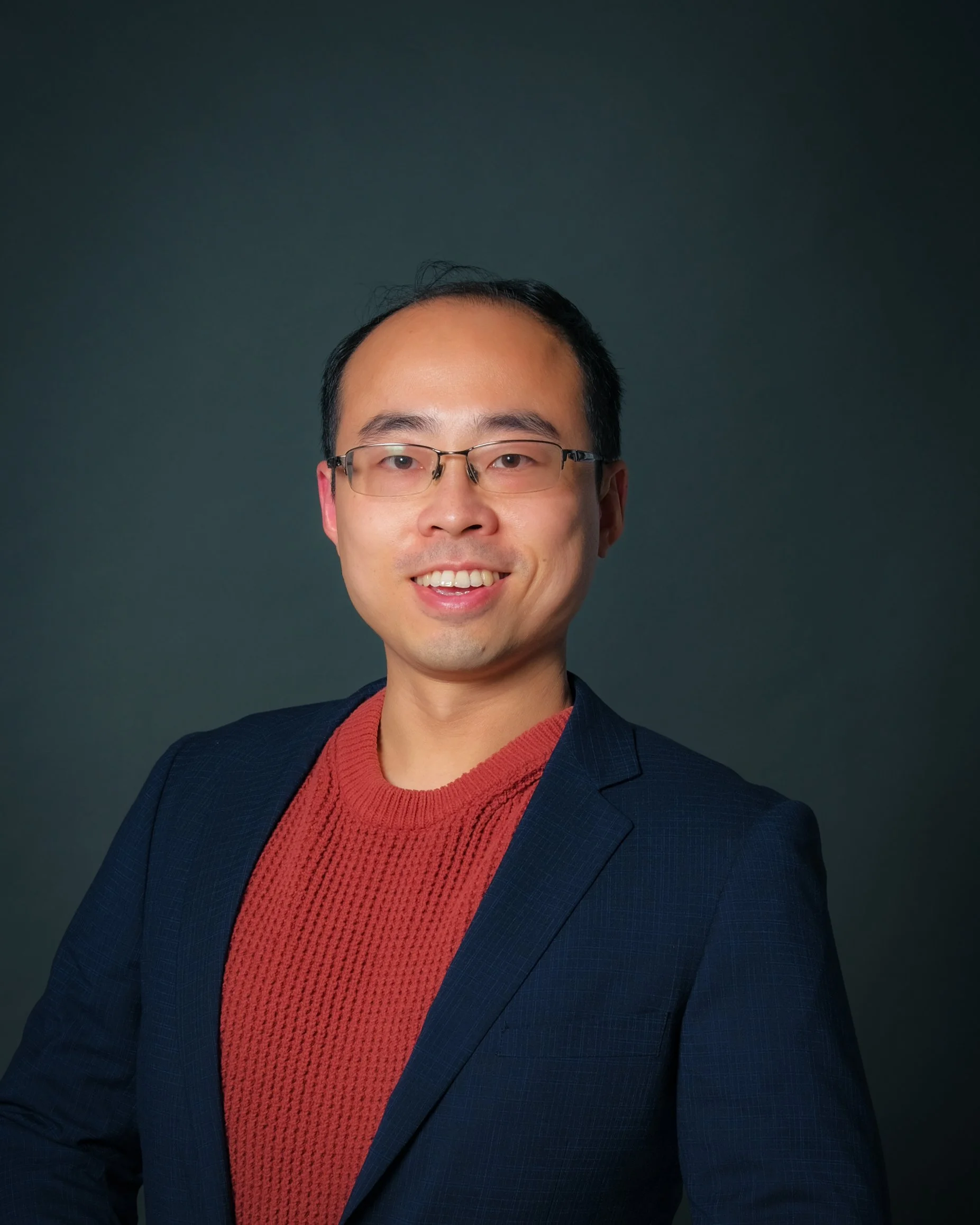 A portrait of a smiling man with glasses, wearing a red sweater and a dark blazer, against a dark background.
