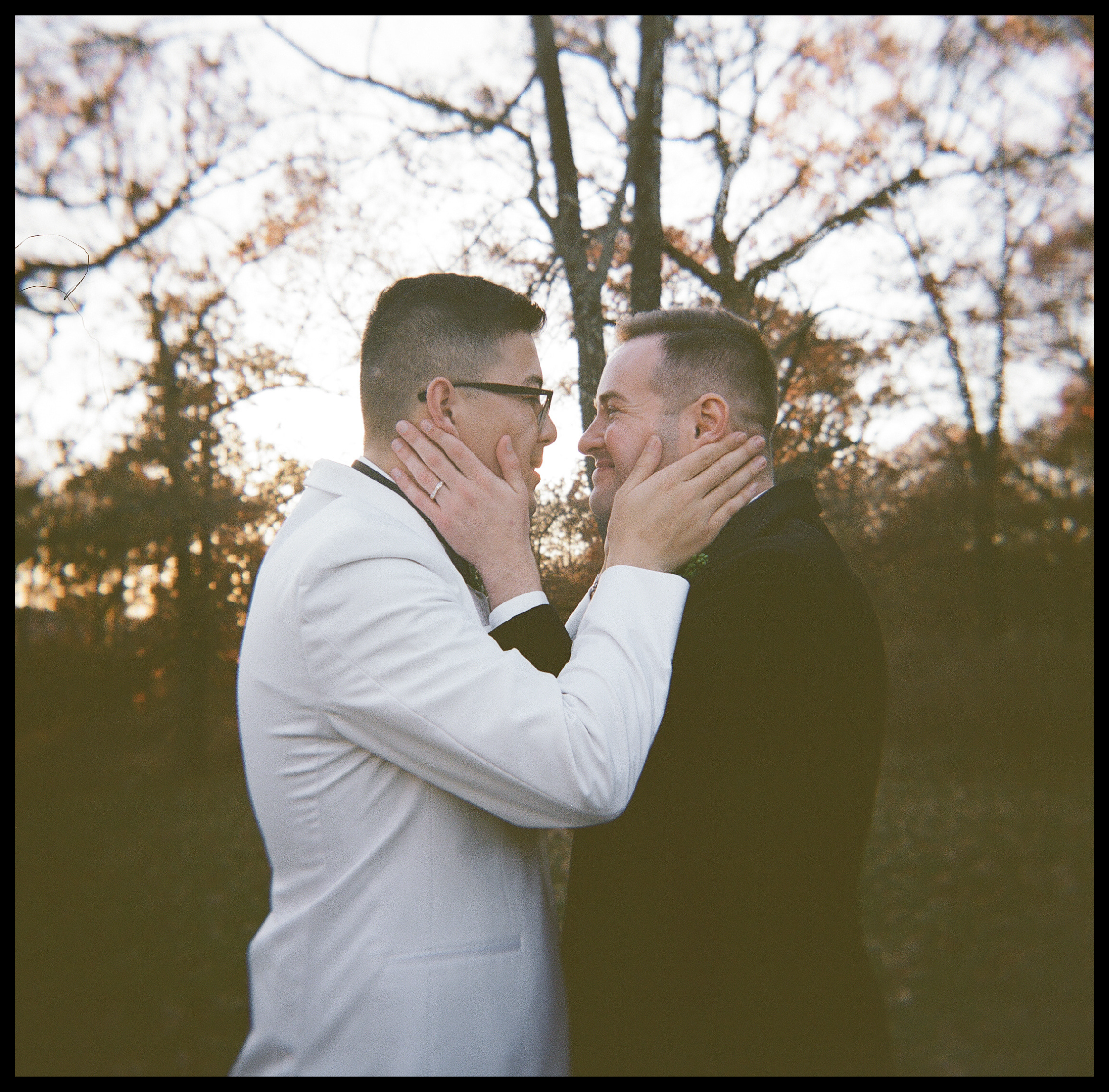 Two men, wearing glasses, are touching each other's faces and looking into each other's eyes with smiles, outdoors with a background of leafless trees and clear sky.