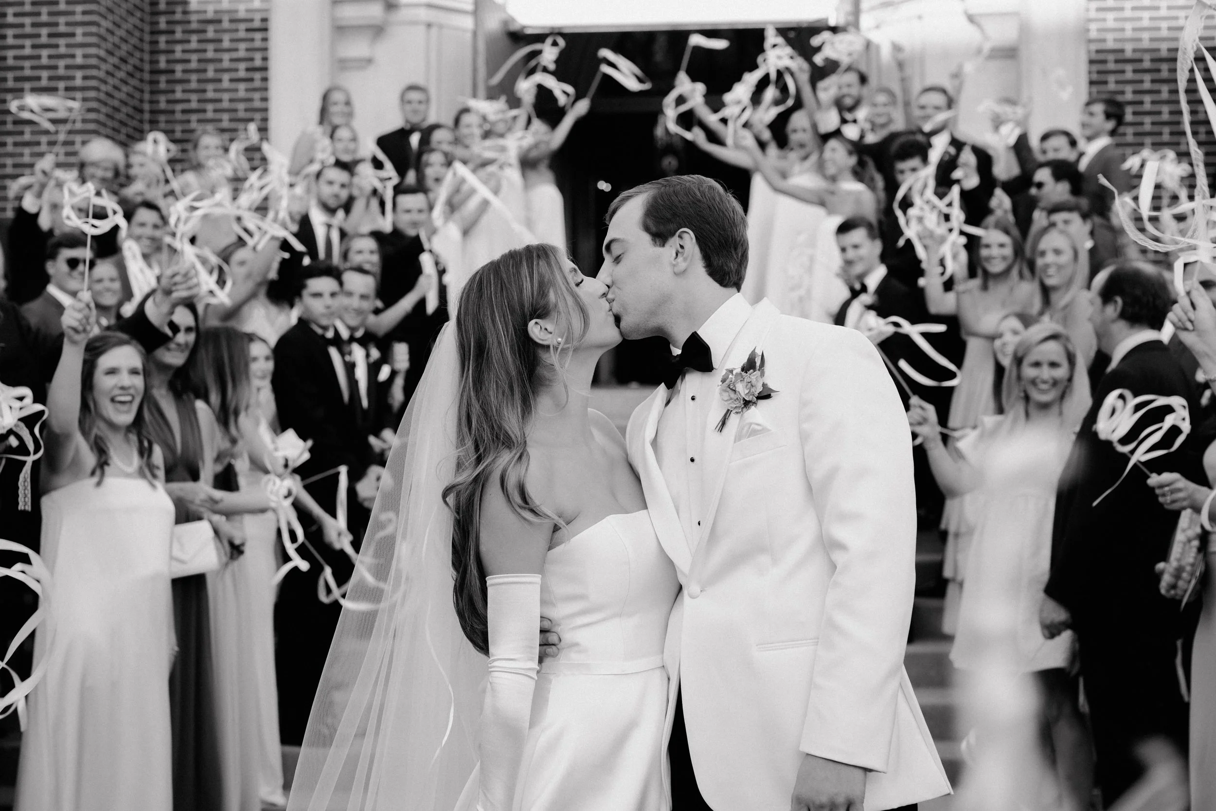 A bride and groom sharing a kiss at their wedding with guests in the background throwing streamers.
