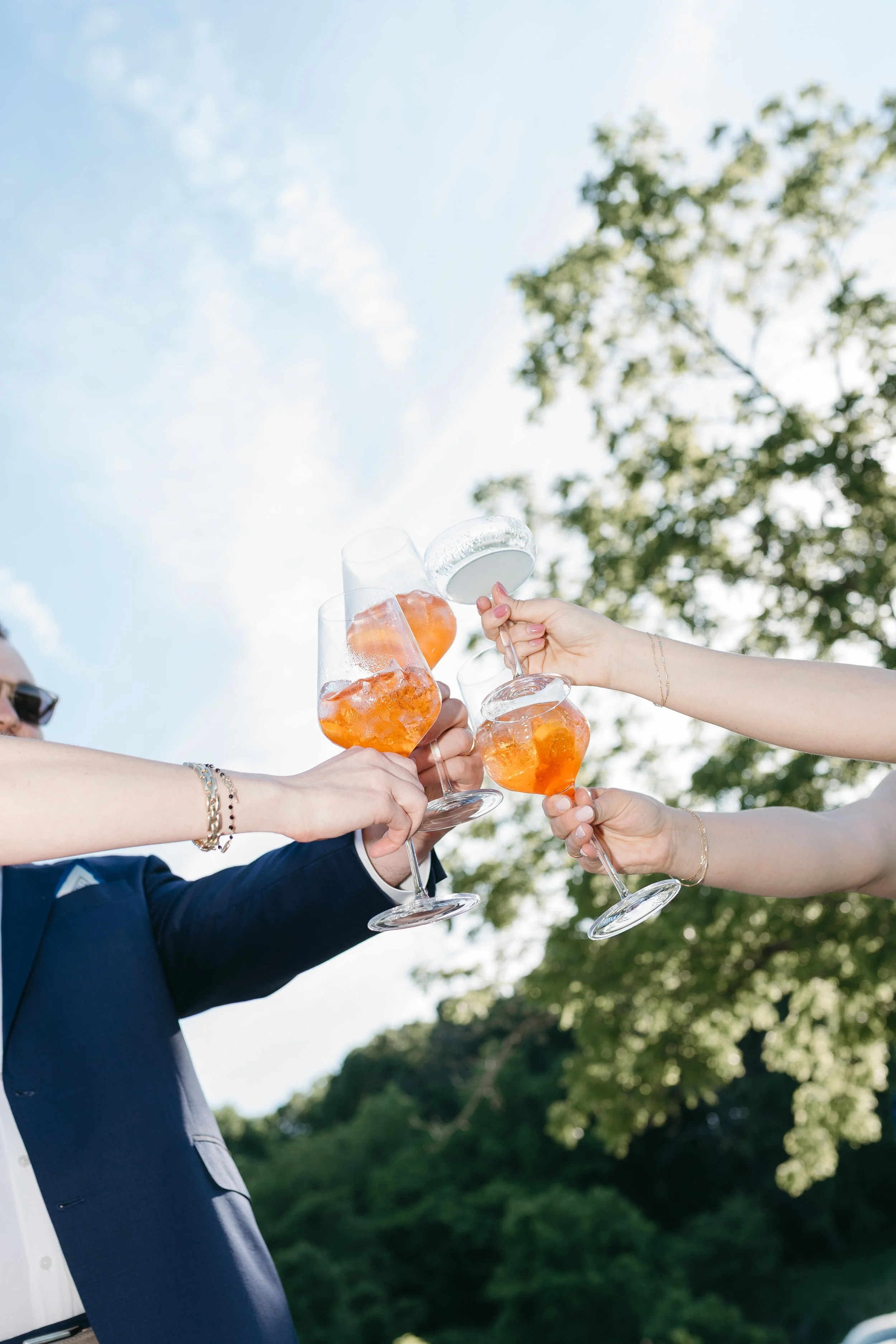 People holding glasses with orange beverage and ice, toasting outdoors on a sunny day, with trees and blue sky in the background.