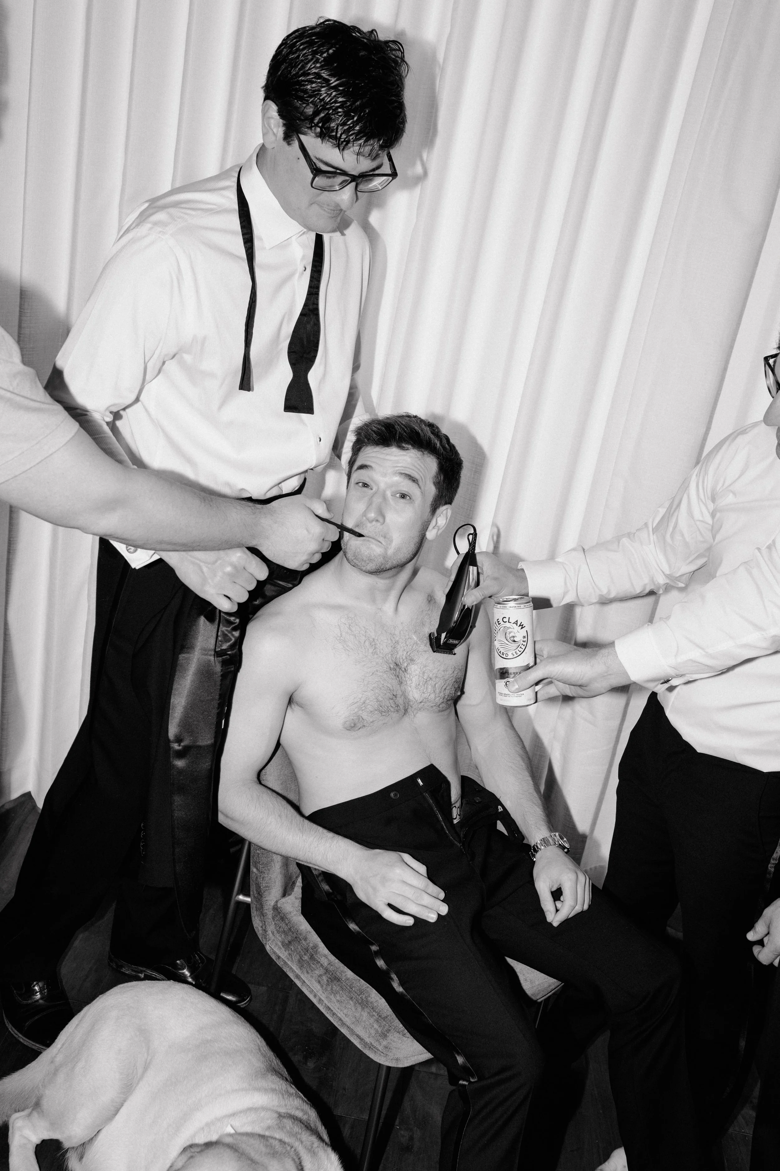 A man is seated shirtless, holding a can of beer, while two men perform tattoos on him. One man is tattooing his face, and the other is holding a tattoo machine near his chest. A dog is lying in the foreground.