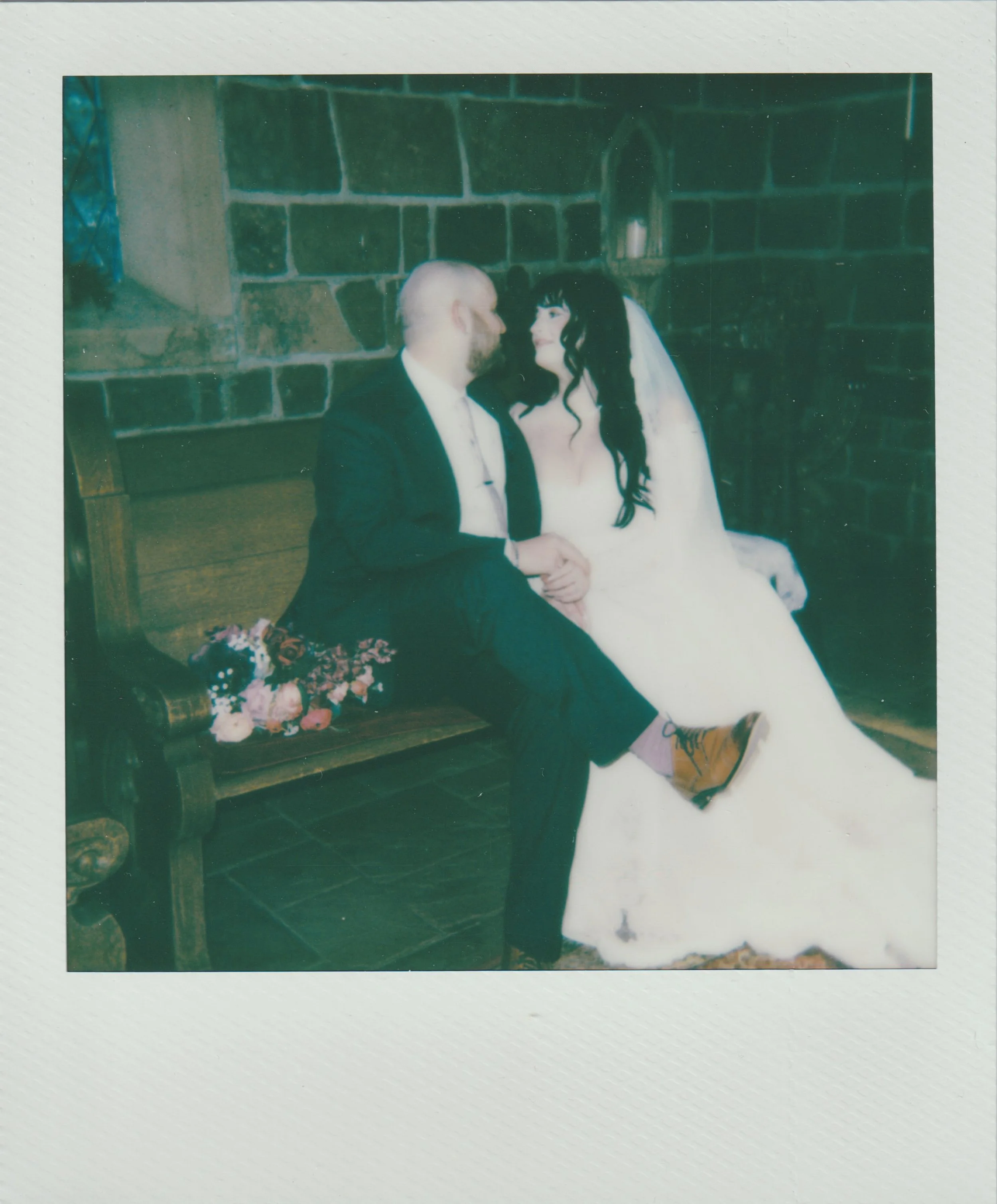 A vintage color photograph of a couple sitting on a wooden bench, gazing into each other's eyes, with a brick wall in the background and flowers nearby.
