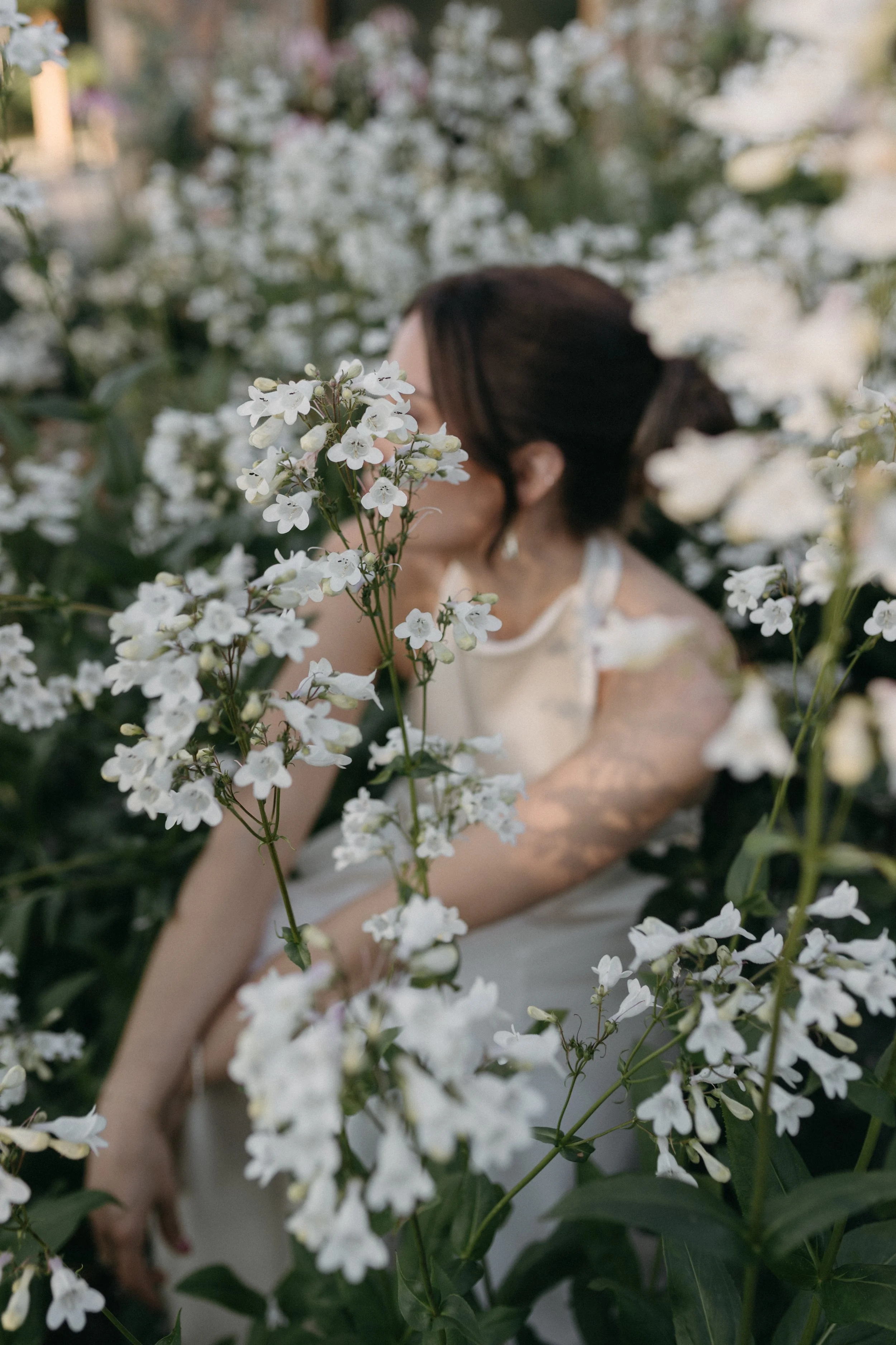 A woman in a white dress surrounded by white flowers, with some flowers she is holding close to her face, in a garden or floral setting.