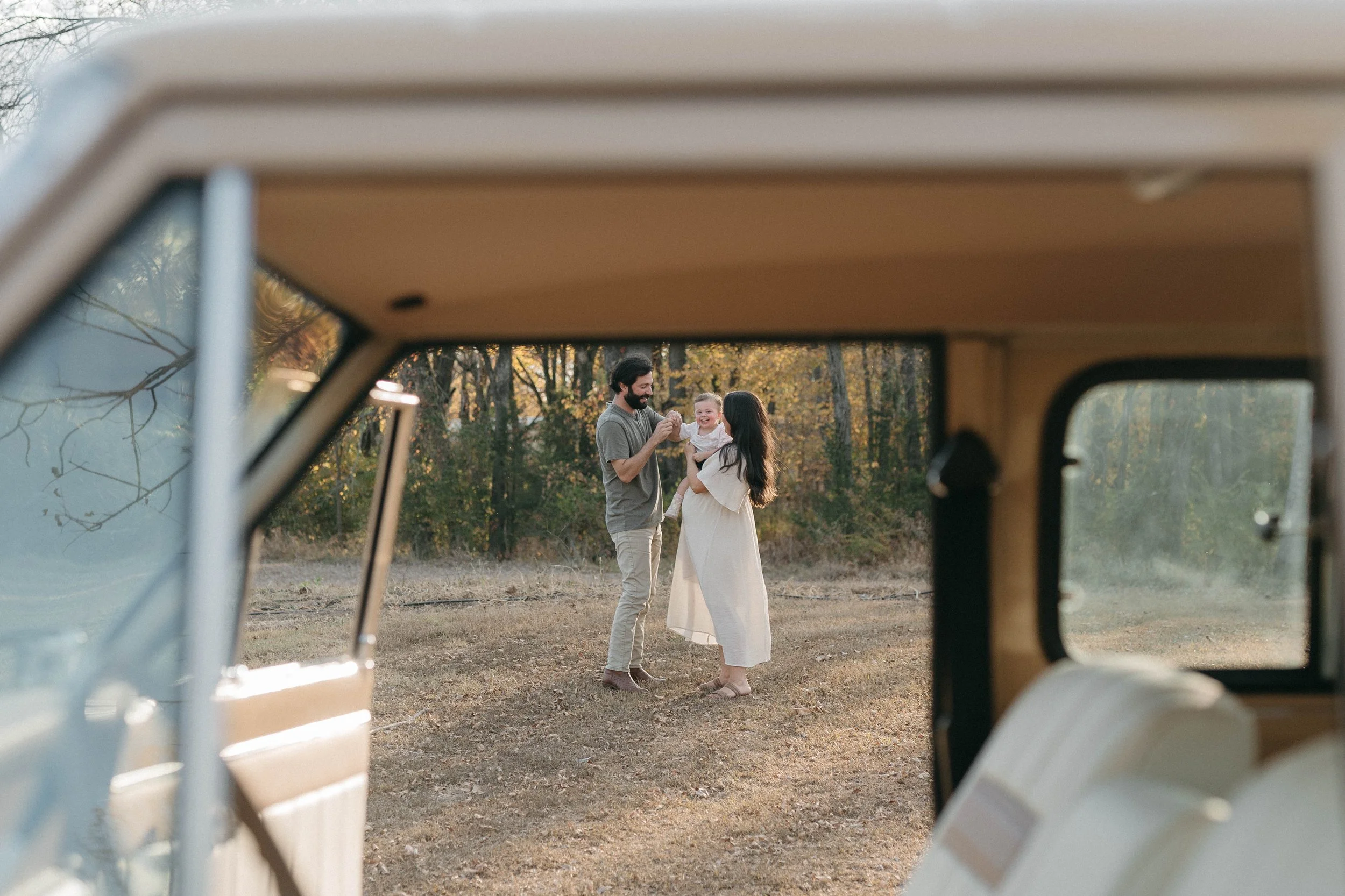 A family of three enjoying time outdoors in a wooded area, seen through the open window of a vintage van.