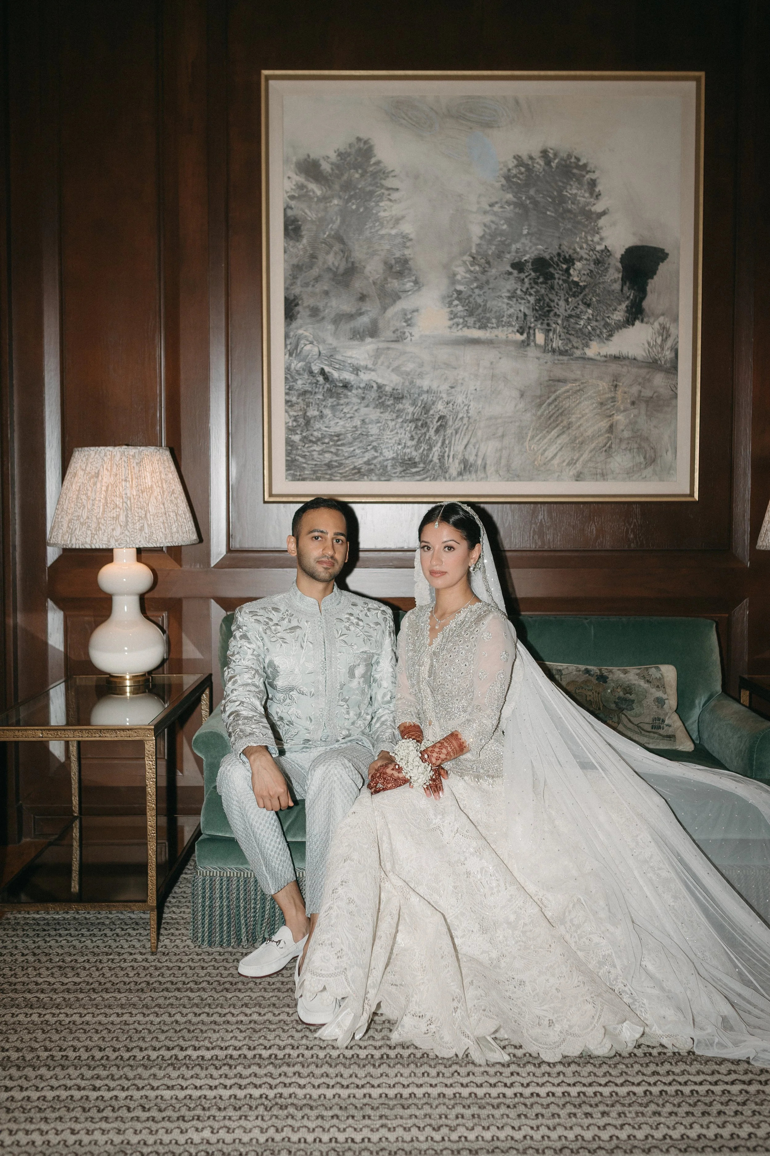 A bride and groom sitting together in a room with dark wood walls, large painting, lamps, and patterned carpet.