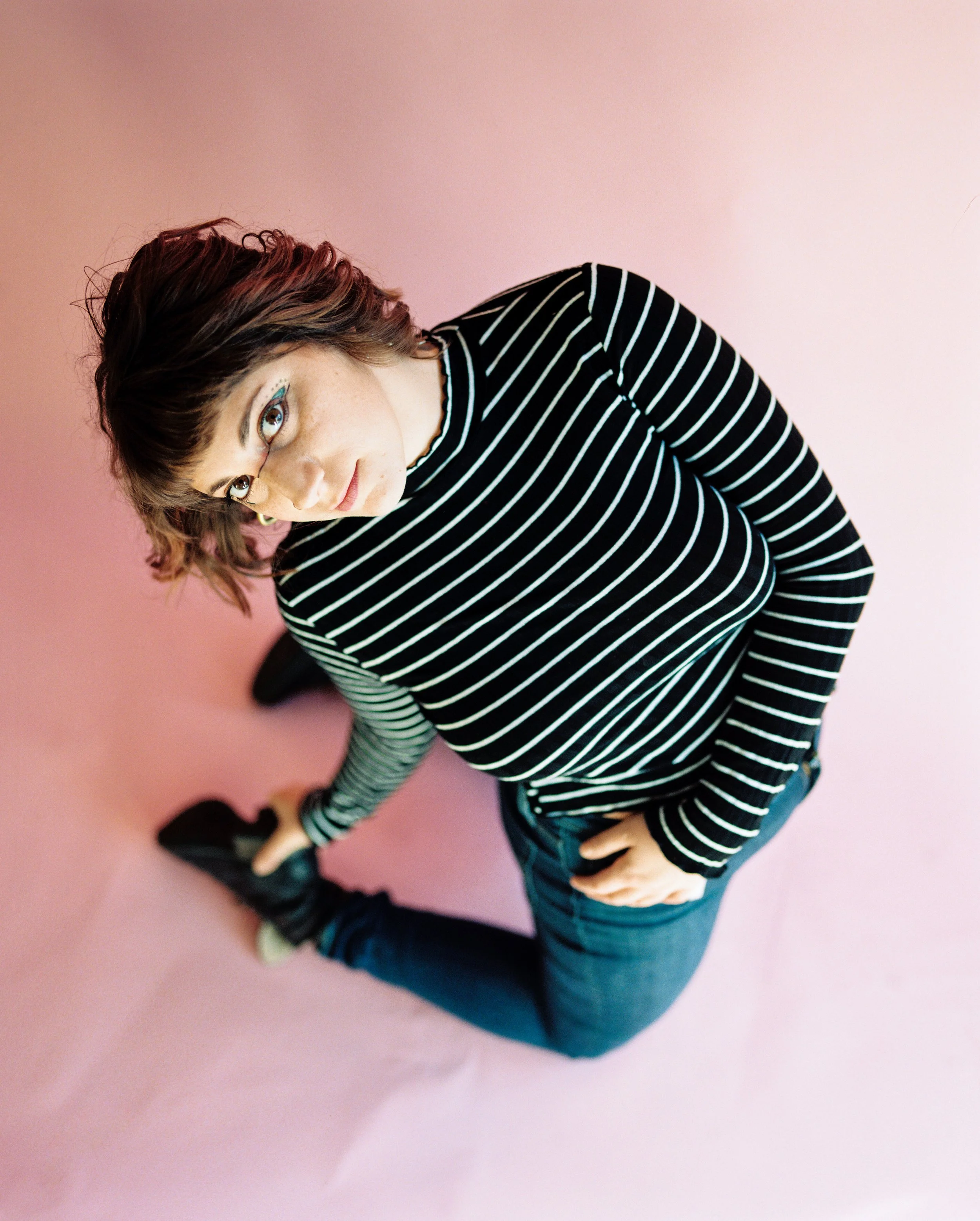 A young woman with shoulder-length hair wearing glasses, a black and white striped long-sleeve shirt, and blue jeans, sitting on a pink background with her legs crossed.
