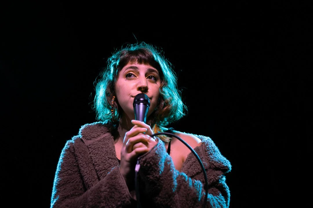 A young woman with short curly hair holding a microphone on stage, wearing a brown fuzzy jacket, illuminated by stage lighting against a dark background.