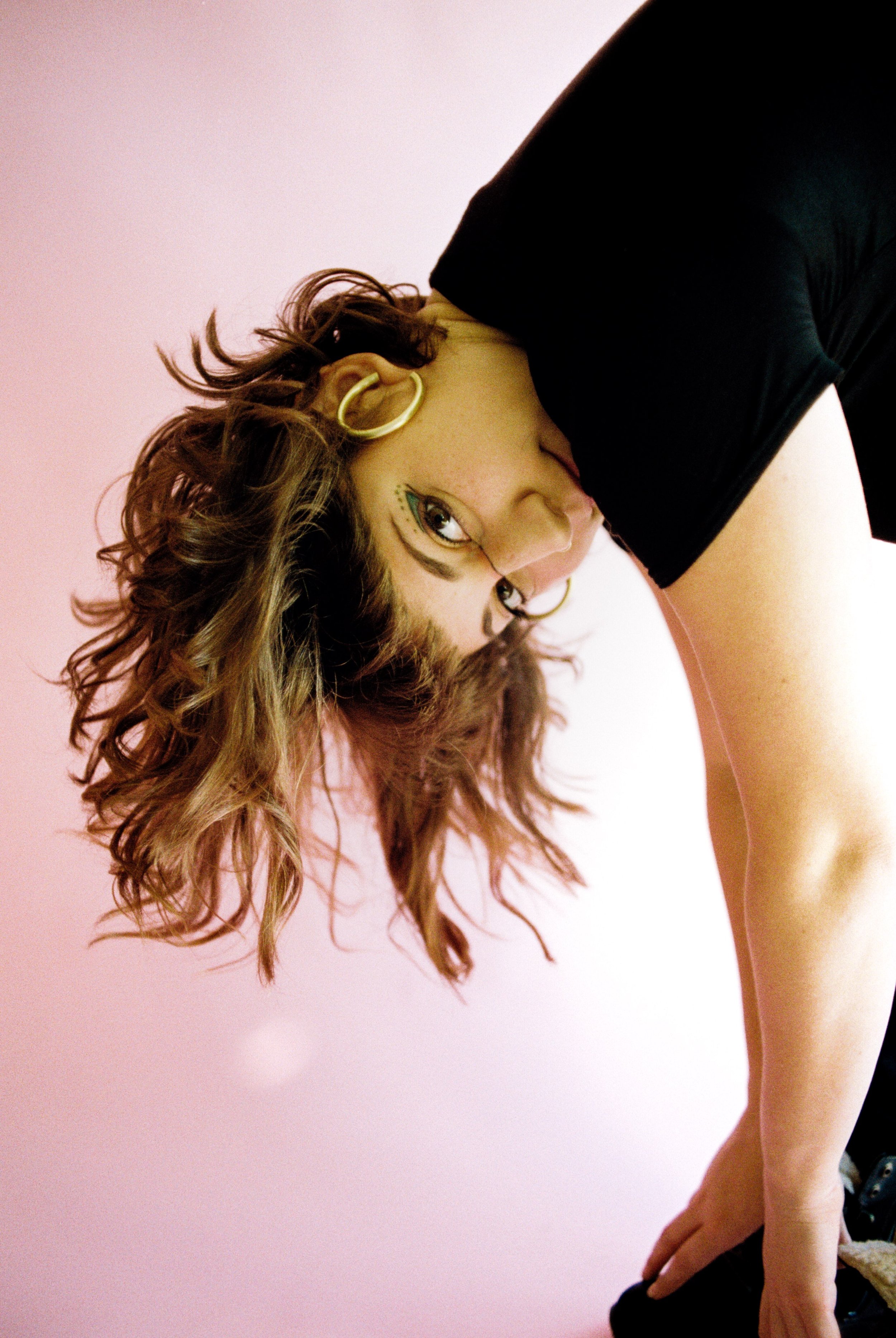A woman with curly hair, makeup, and hoop earrings leaning over and touching the floor, with a pink background.