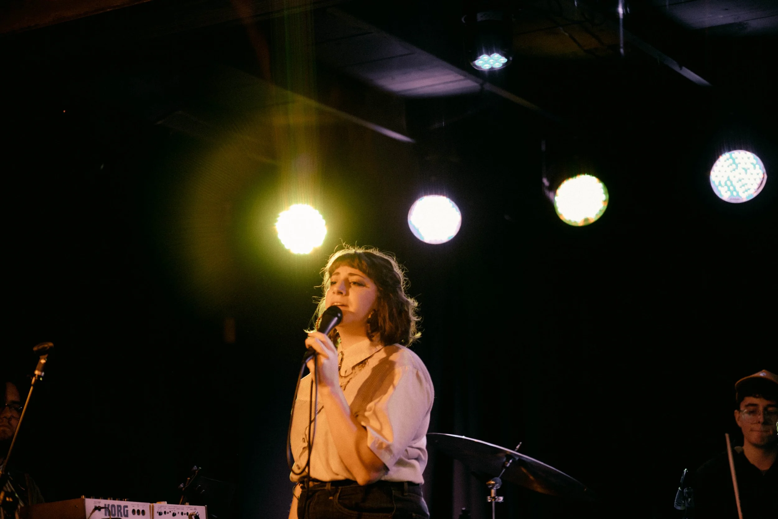 A young woman with short brown hair holding a microphone on stage, with colorful stage lights overhead, partially visible musicians, and a keyboard named Korg in the foreground.