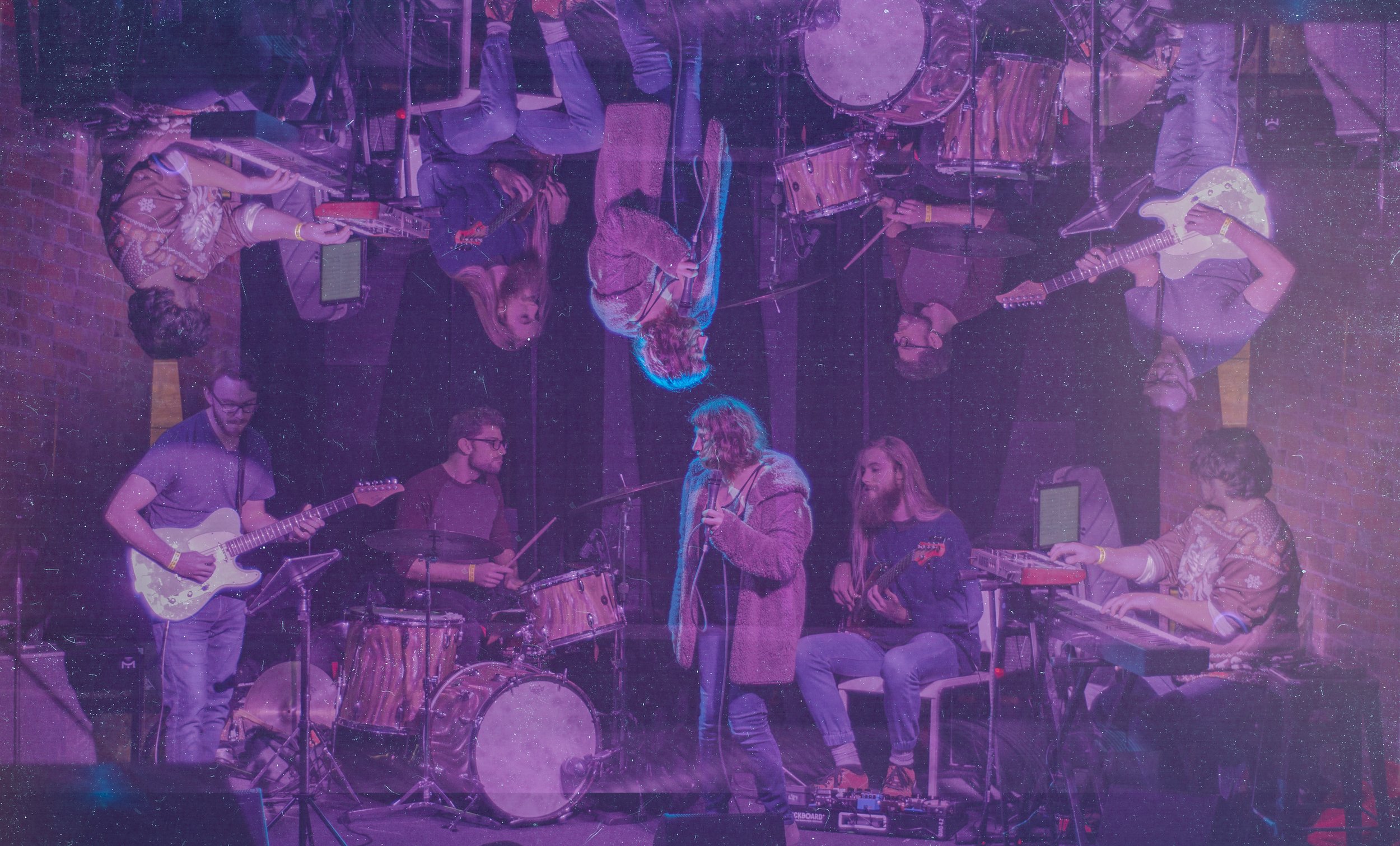 A group of musicians performing on stage with a purple and pink lighting, including guitarists, drummers, and singers, in an indoor concert setting.